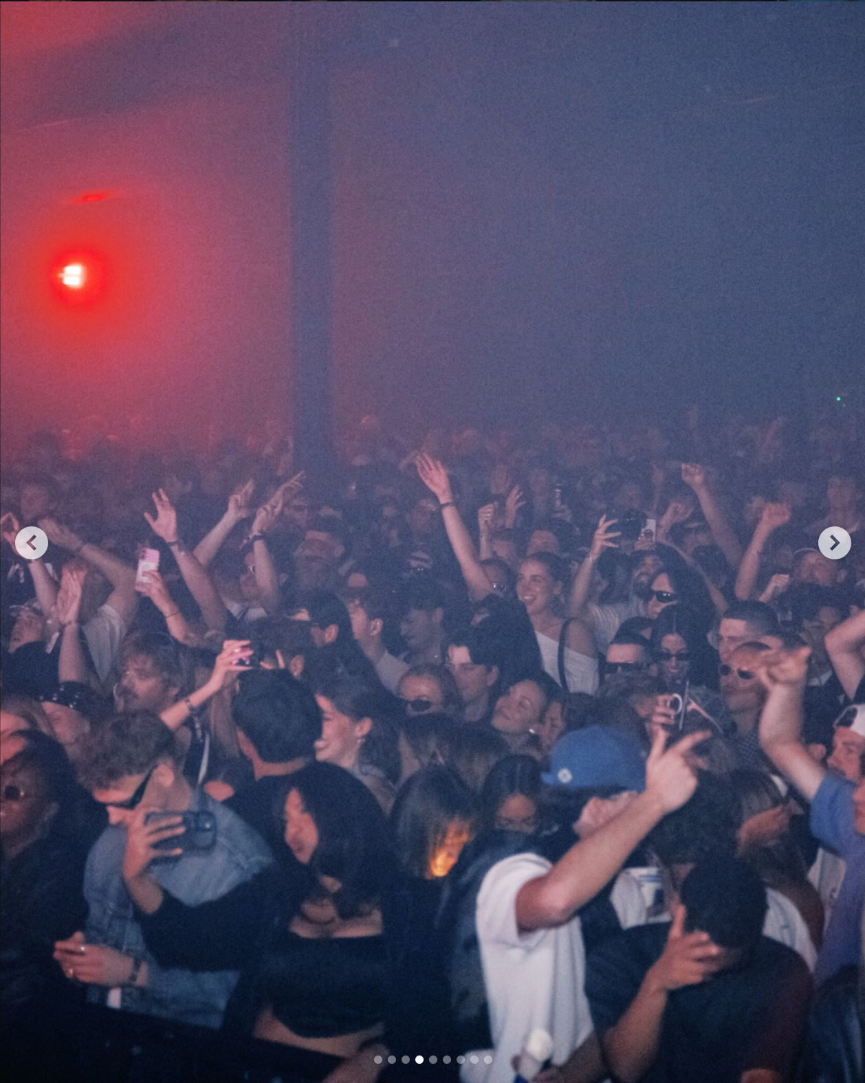 A large crowd of people at a concert or nightclub dancing and taking photos with their phones under dim colorful lighting.
