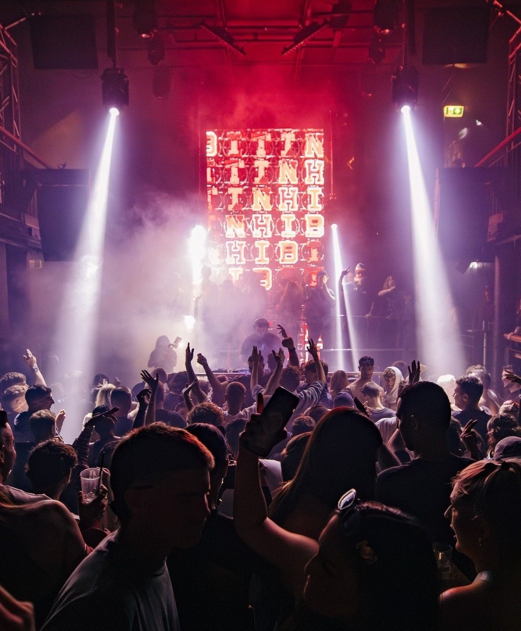 People dancing and enjoying a concert or nightclub with colorful lights, smoke effects, and a large screen displaying illuminated text in the background.