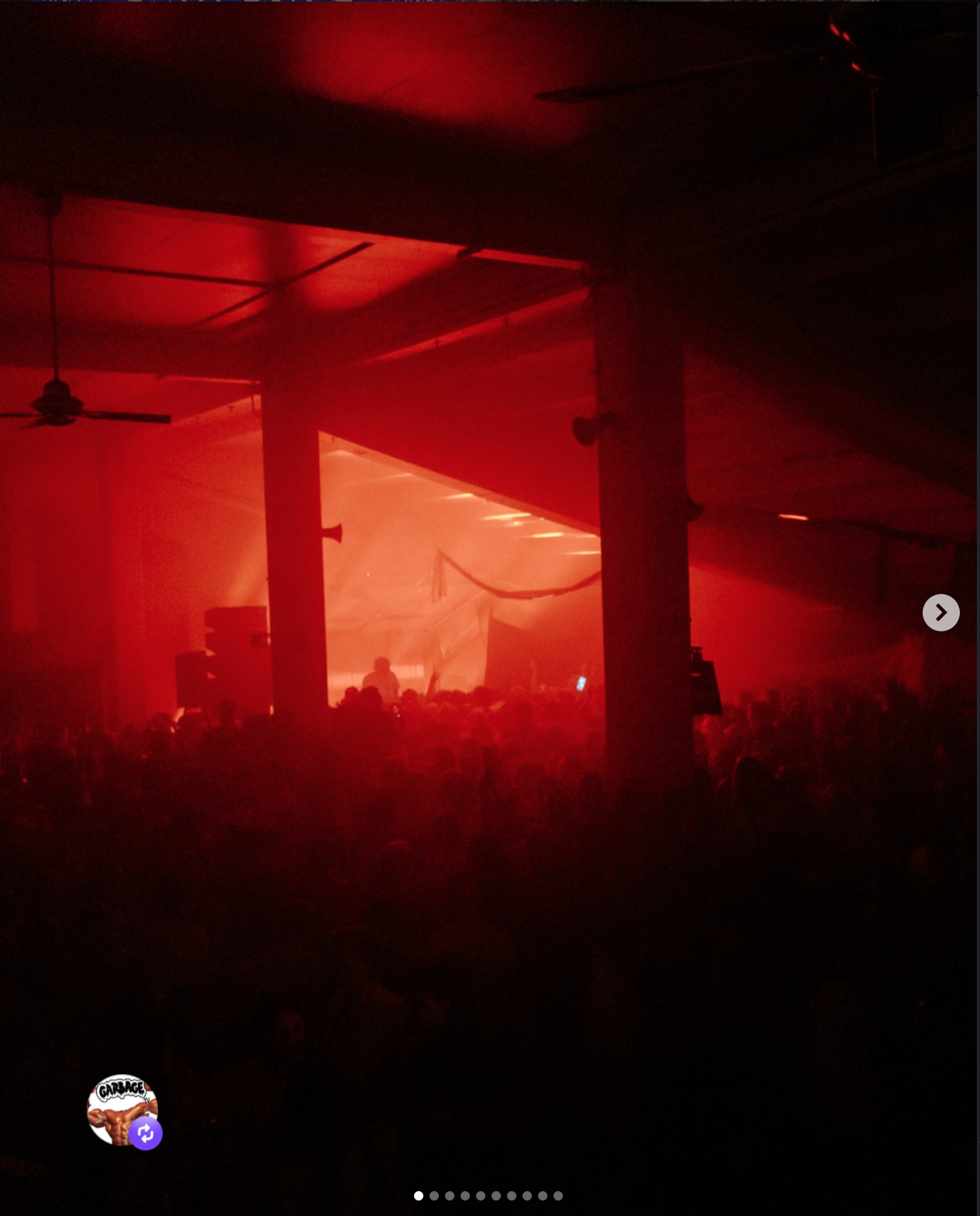 A dark indoor concert venue illuminated by red lighting, with silhouettes of a crowd and a DJ or performer on stage in the background.