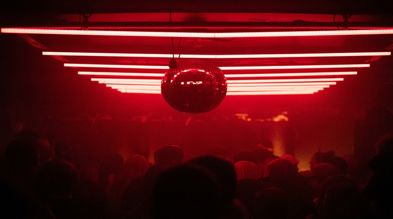A dark dance club with red neon lighting, a disco ball hanging from the ceiling, and silhouettes of people dancing or standing, facing the stage or DJ area.