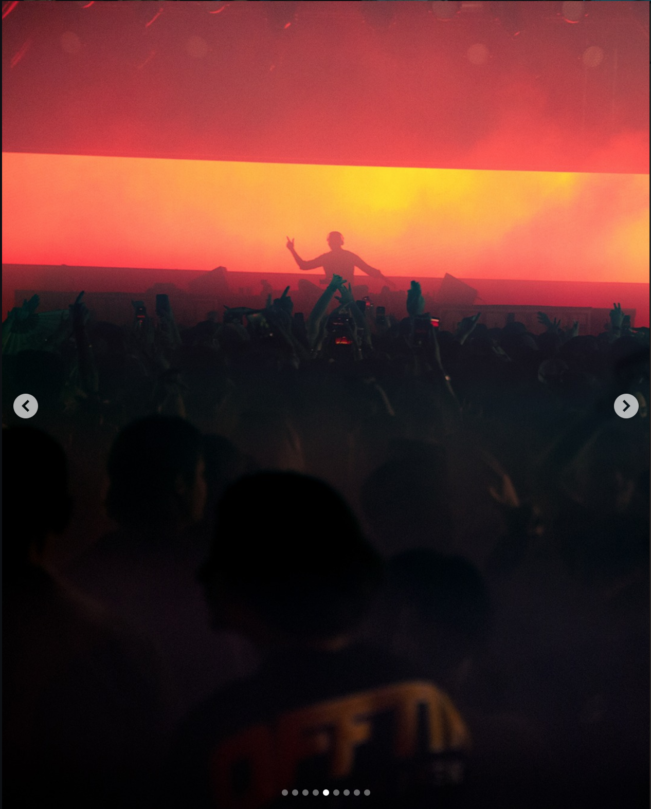 A person on stage making a peace sign with a crowd raising hands at a concert or event with a large screen displaying a colorful sunset or abstract background.