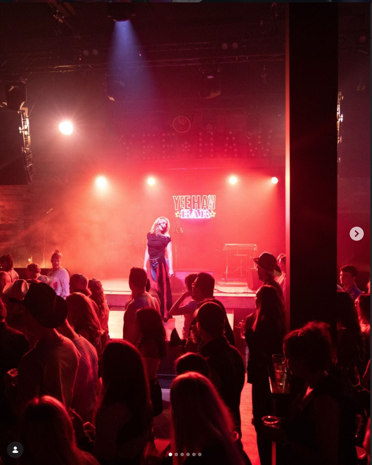 A stage with a performer in front of a microphone, illuminated by red and purple stage lights. The audience is seated at tables, watching the performance. A sign in the background reads 'YEEHAW BAR'.