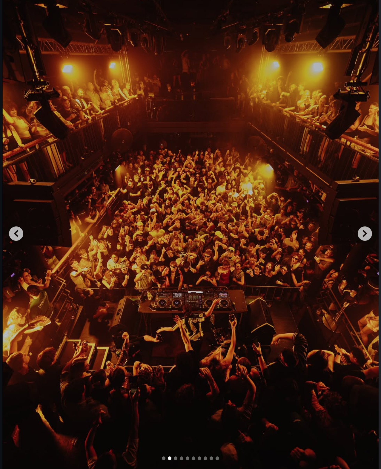 Crowd dancing and enjoying music at a nightclub with DJ booth, yellow lighting, and two levels of balconies.