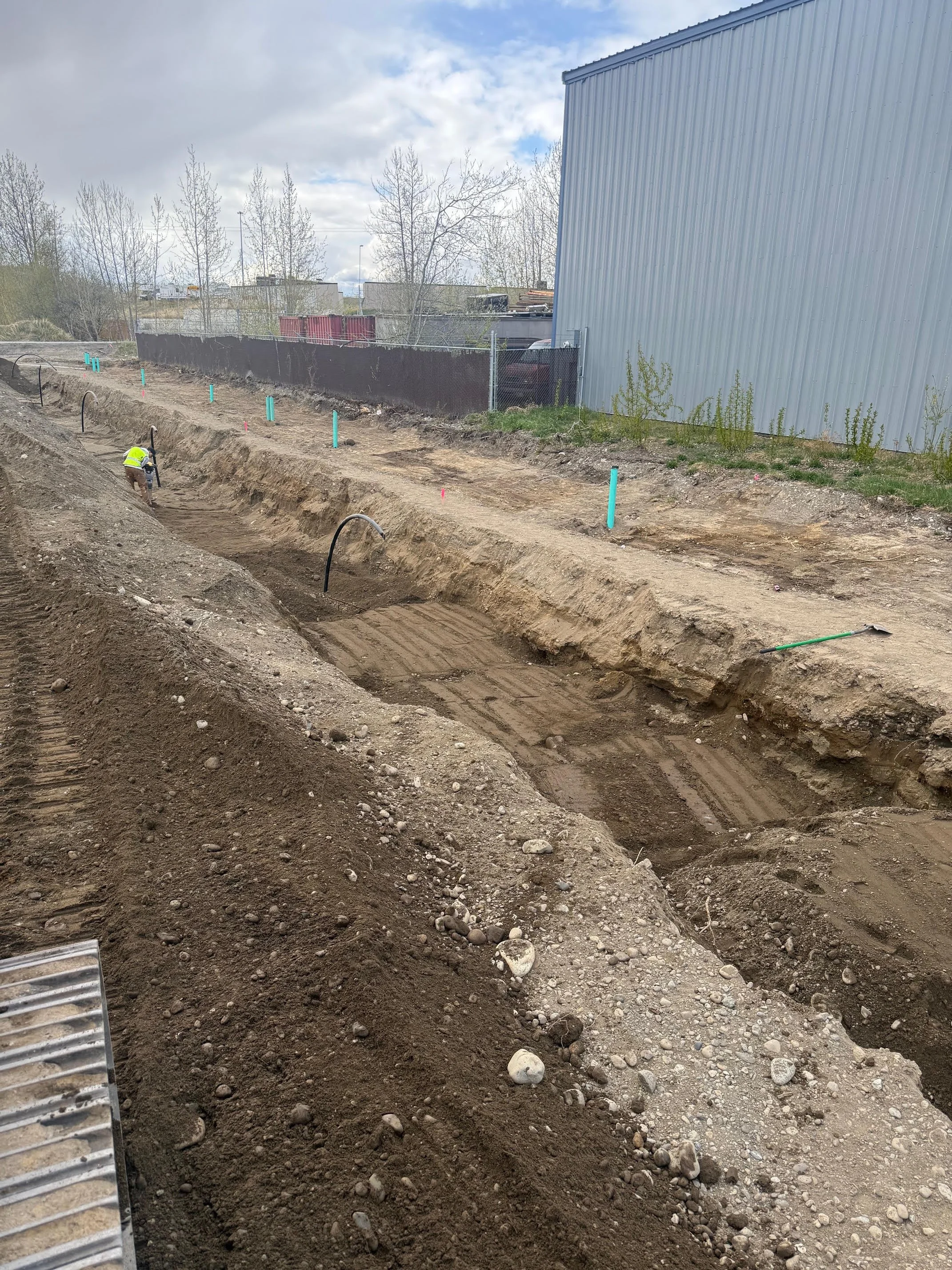 Construction site with excavated trenches, a worker in a yellow safety vest, and construction pipes sticking out of the ground.