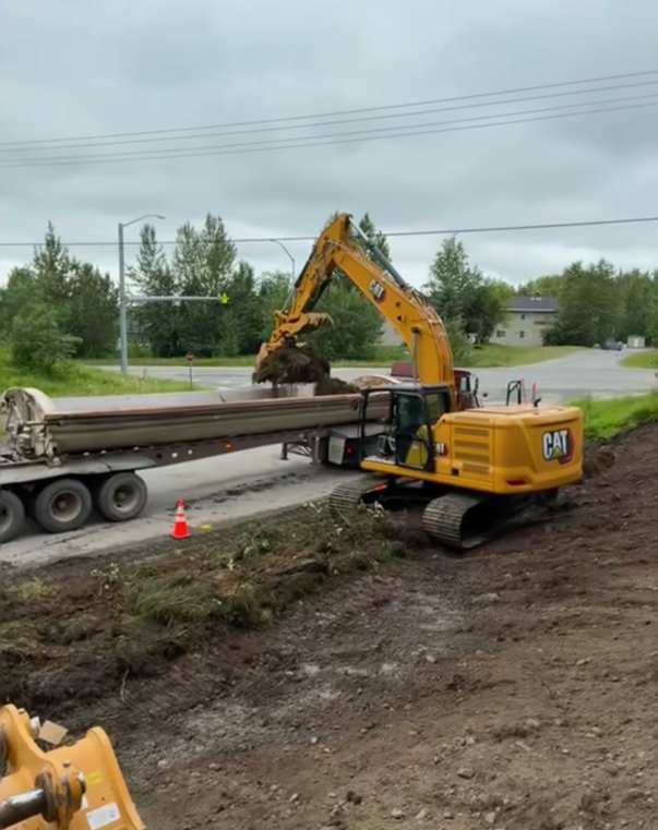 A yellow CAT excavator loading soil into a semi-truck on a construction site near a road.
