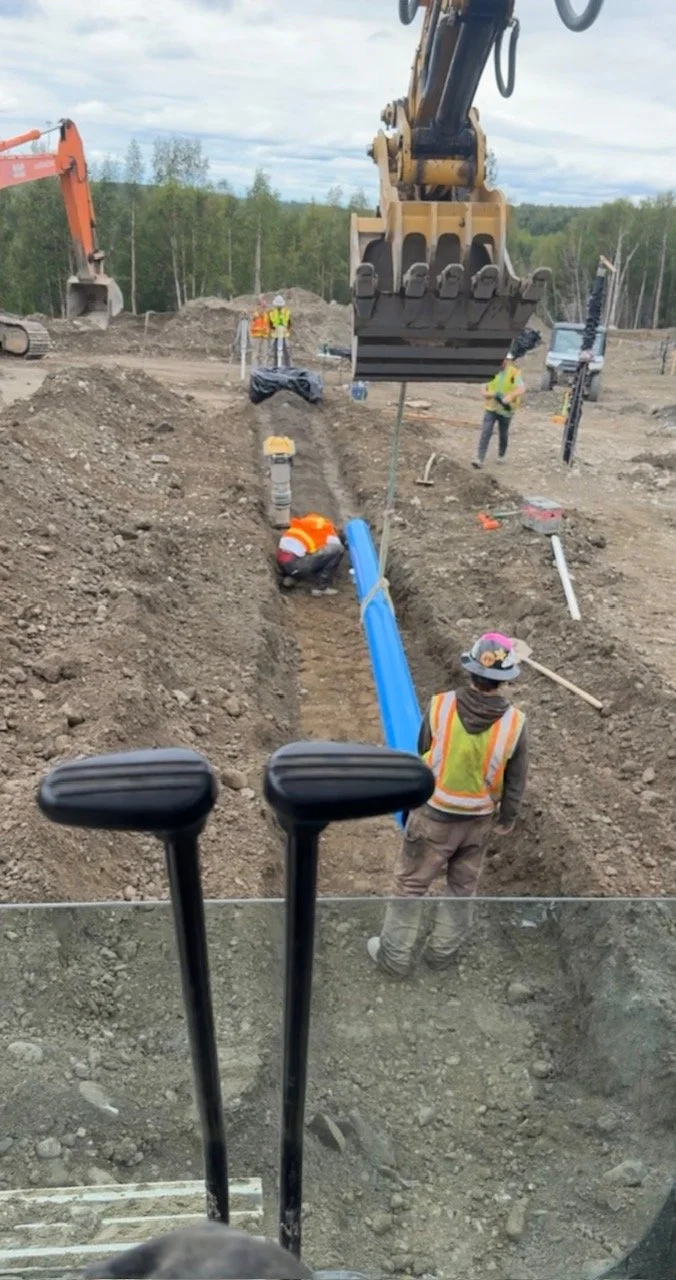 Construction site with workers installing large blue underground pipes, excavated trench, and heavy machinery including an excavator and a backhoe.