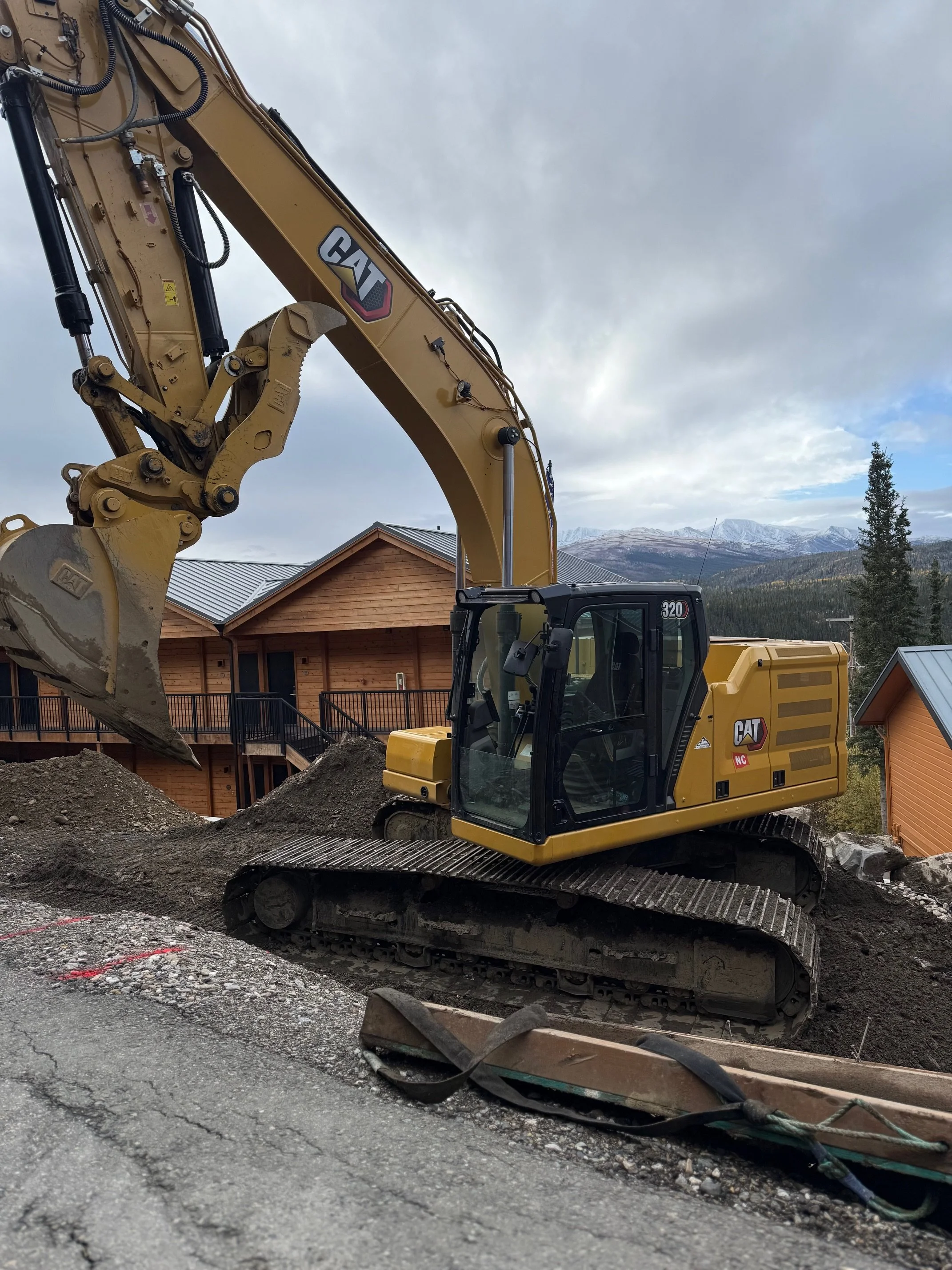 A yellow CAT excavator working on a dirt construction site in a mountainous area with wooden houses in the background and cloudy sky.