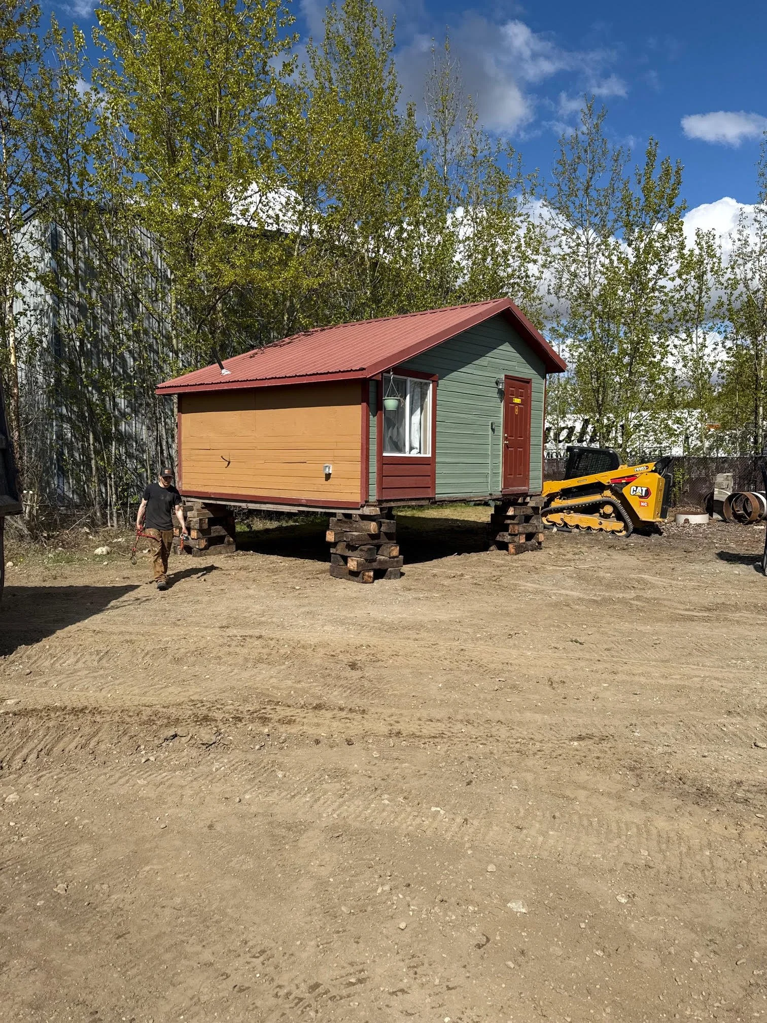A tiny house on stilts with a red roof, green and yellow walls, and a window, situated in a yard with trees and construction equipment nearby.