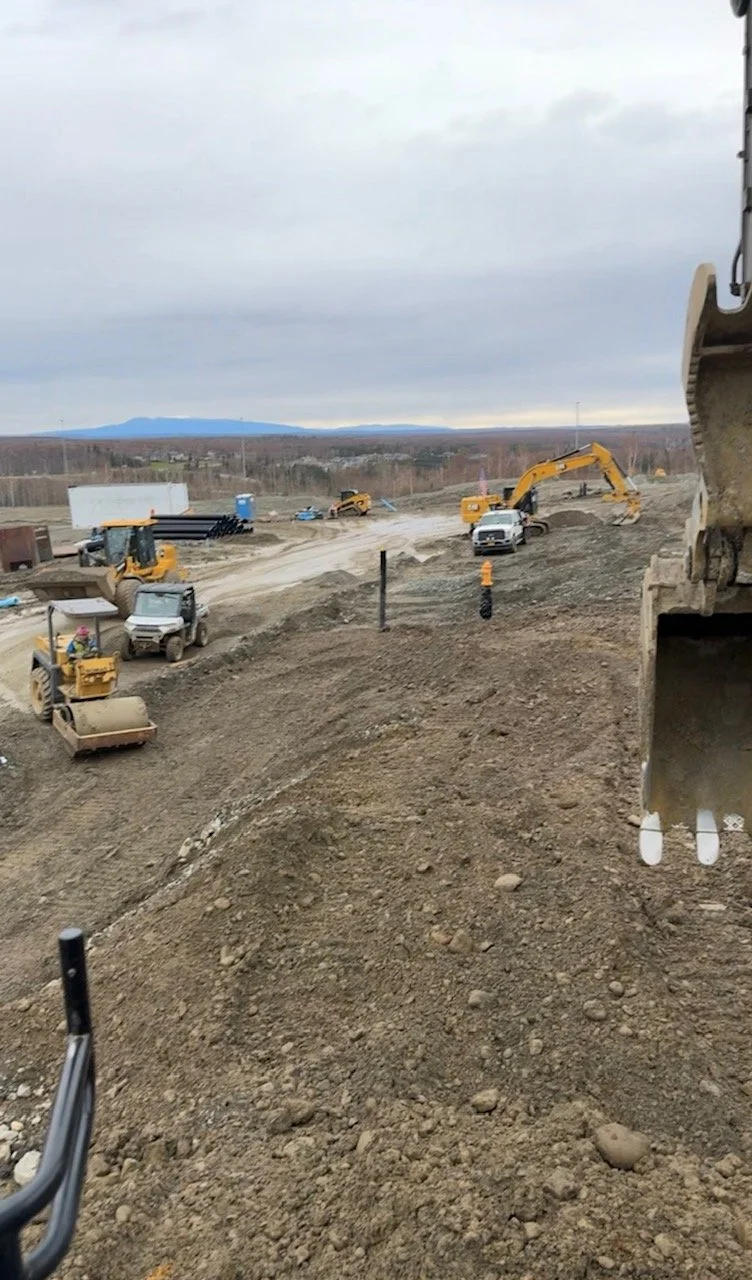 Construction site with heavy machinery, including excavators, bulldozers, and trucks, working on the dirt terrain under a cloudy sky.