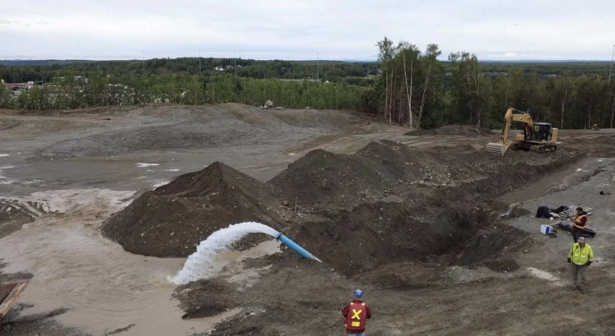 Construction site with a yellow excavator, piles of dirt, and workers near water pipe.