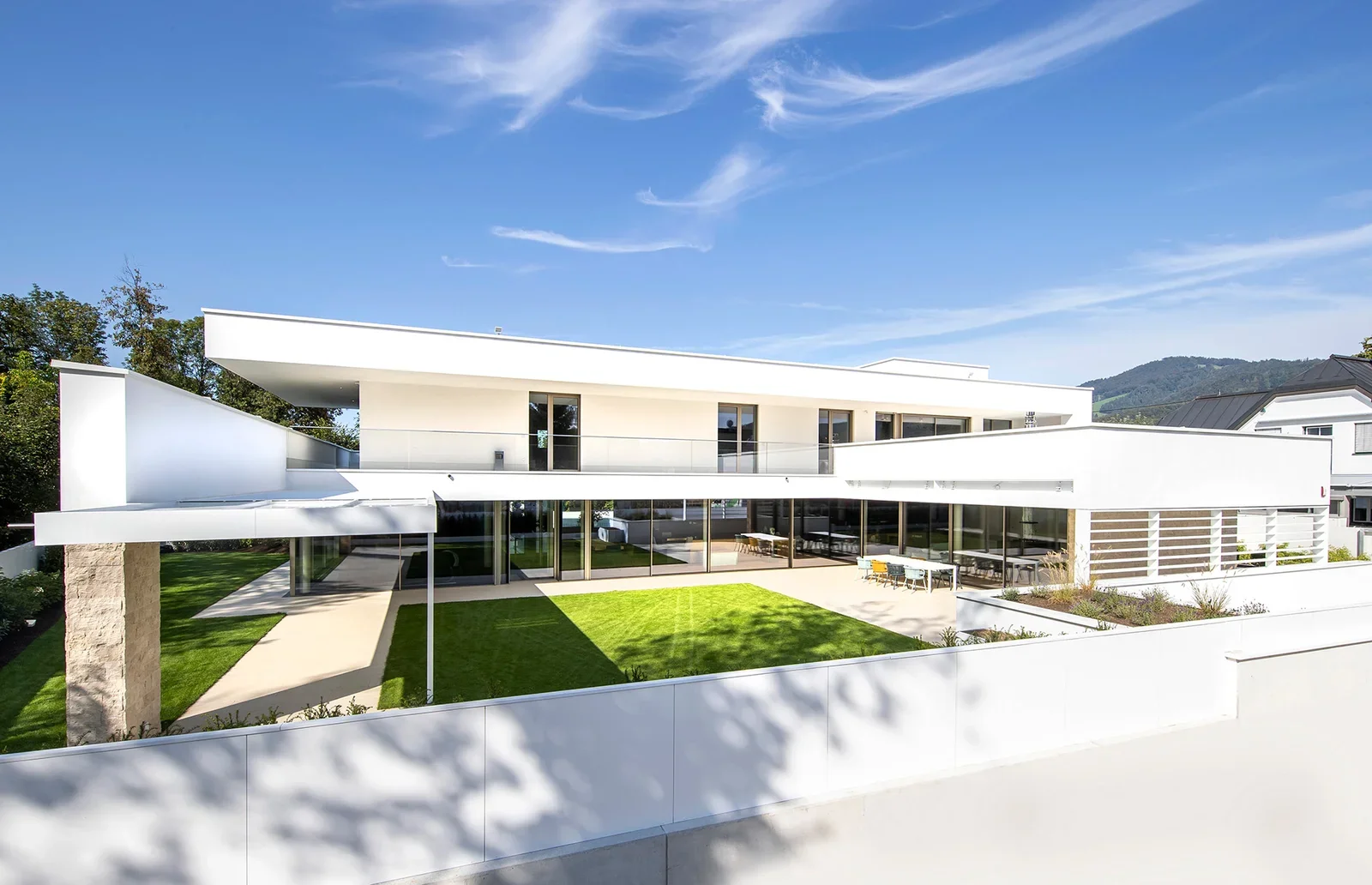 Modern white multi-story house with large glass windows, a well-manicured lawn, outdoor patio, and a backdrop of trees and mountains under a clear blue sky.