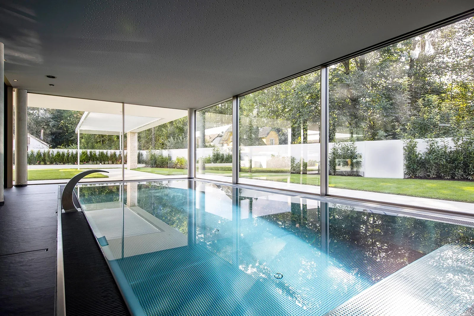 Indoor swimming pool with large glass windows showing a green backyard with trees and house in the background.