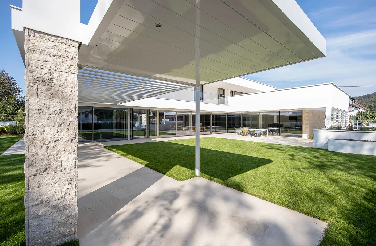 Modern house with large glass windows, white walls, and a covered patio area with green grass and a concrete walkway.