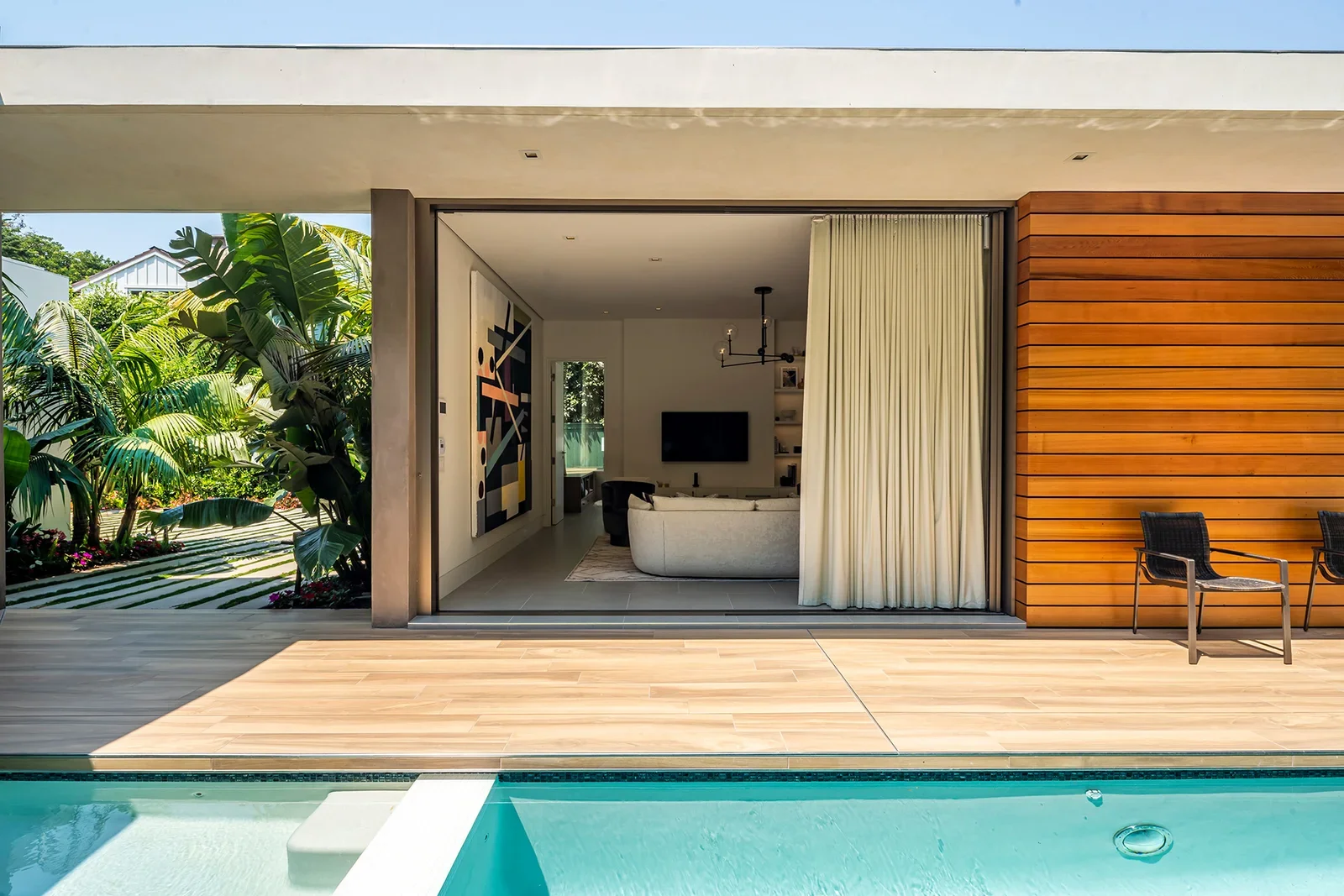 Modern house with an open sliding door leading to a living room, wooden deck, outdoor seating, and a pool in the foreground, surrounded by green plants.