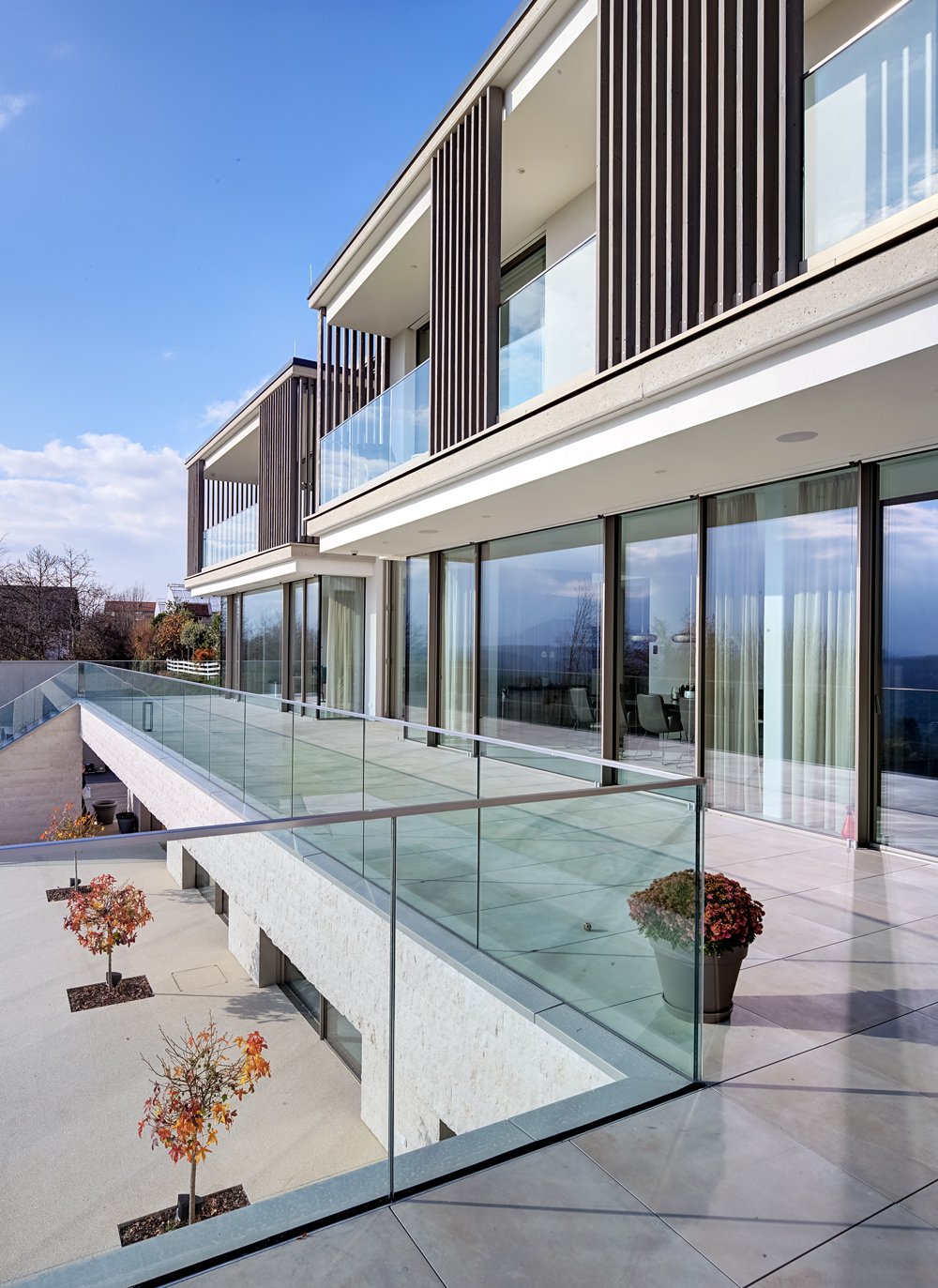 Modern apartment building with large glass windows and balconies, glass railing on the balcony, outdoor potted plants, and a clear sky.