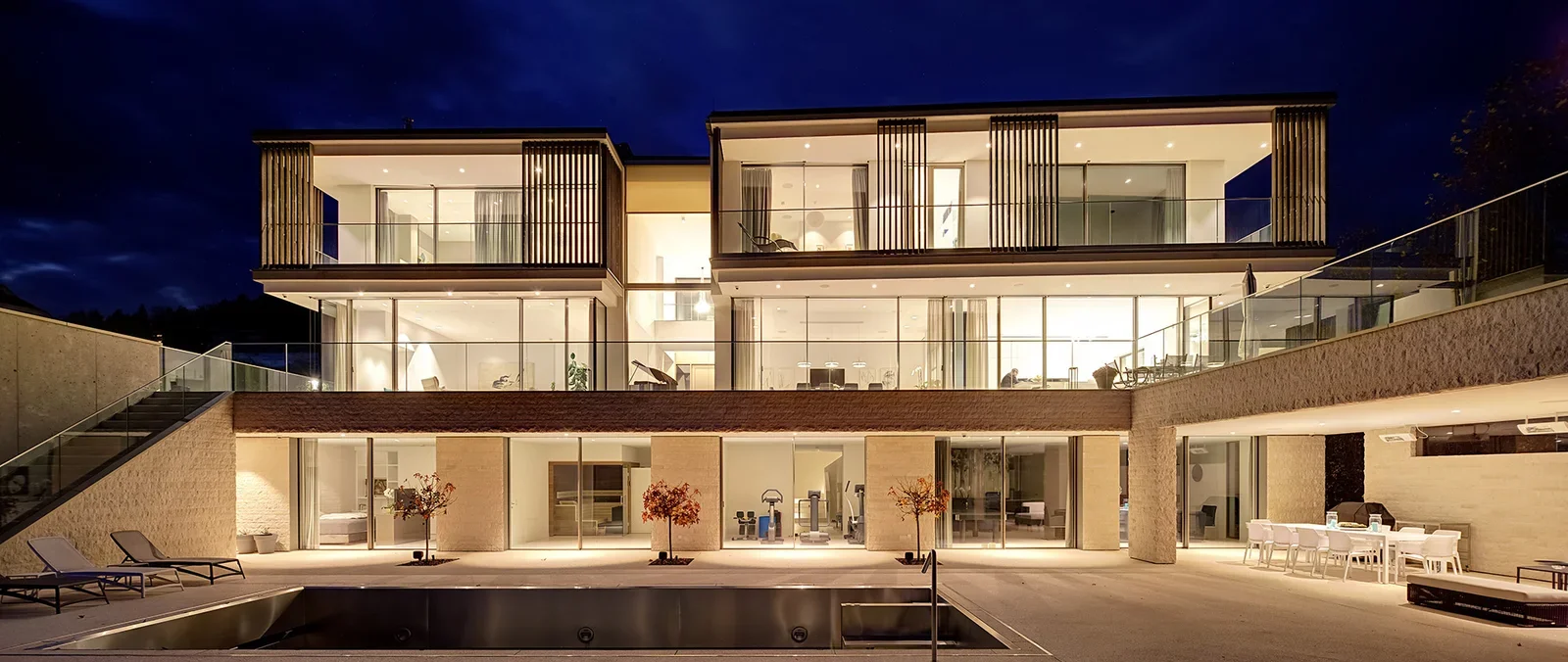 Modern multi-story house at night with large glass windows, interior lighting, outdoor seating area, swimming pool, and landscaped yard.