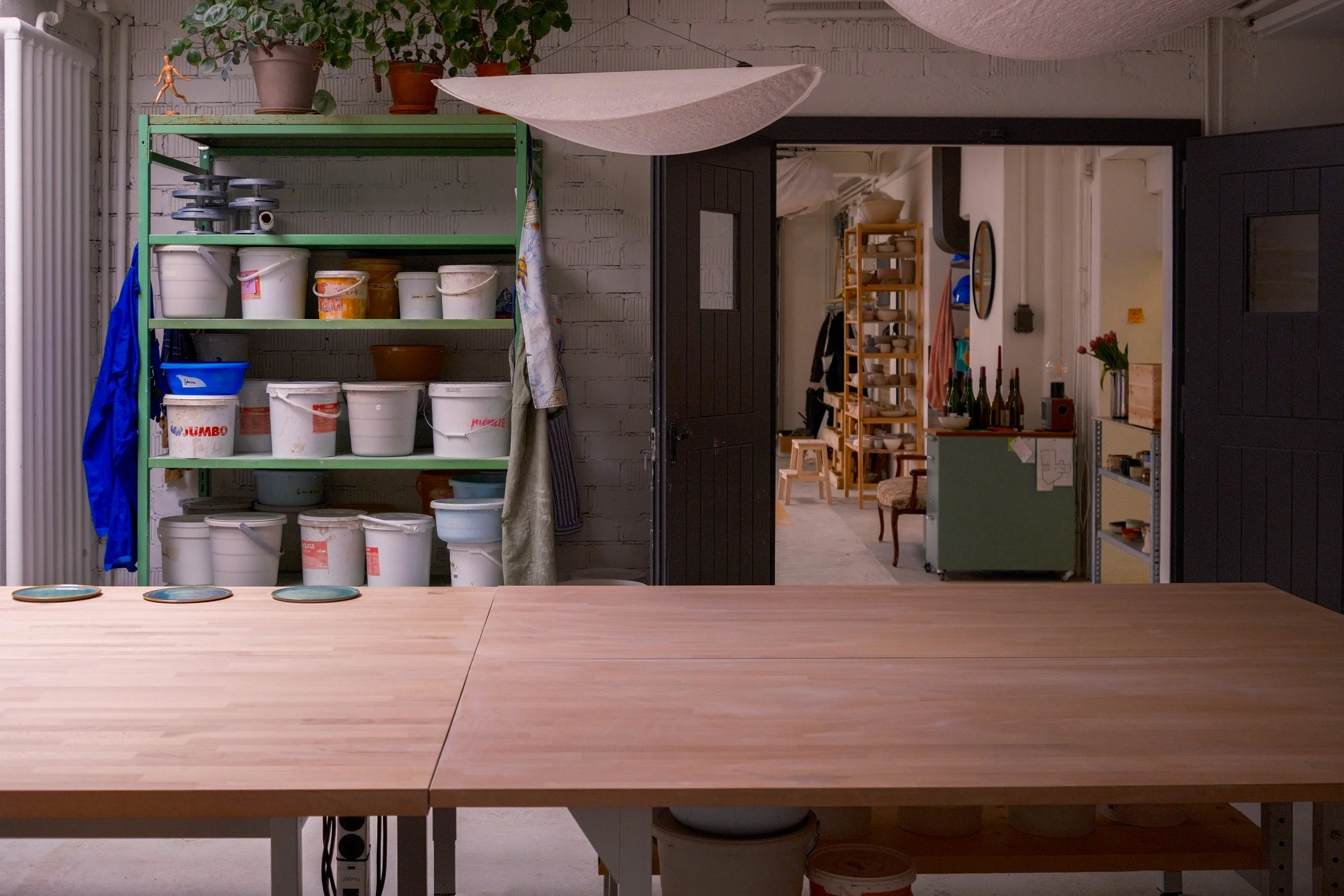 Ein Blick in eine kreative Werkstatt mit einem großen leeren Holztisch im Vordergrund. Im Hintergrund sind offene Regale mit Farben, Töpferwaren und Werkzeugen, sowie eine Küche mit Töpfen und Flaschen. Die Werkstatt hat eine industrielle Atmosphäre mit weißen Ziegelwänden.