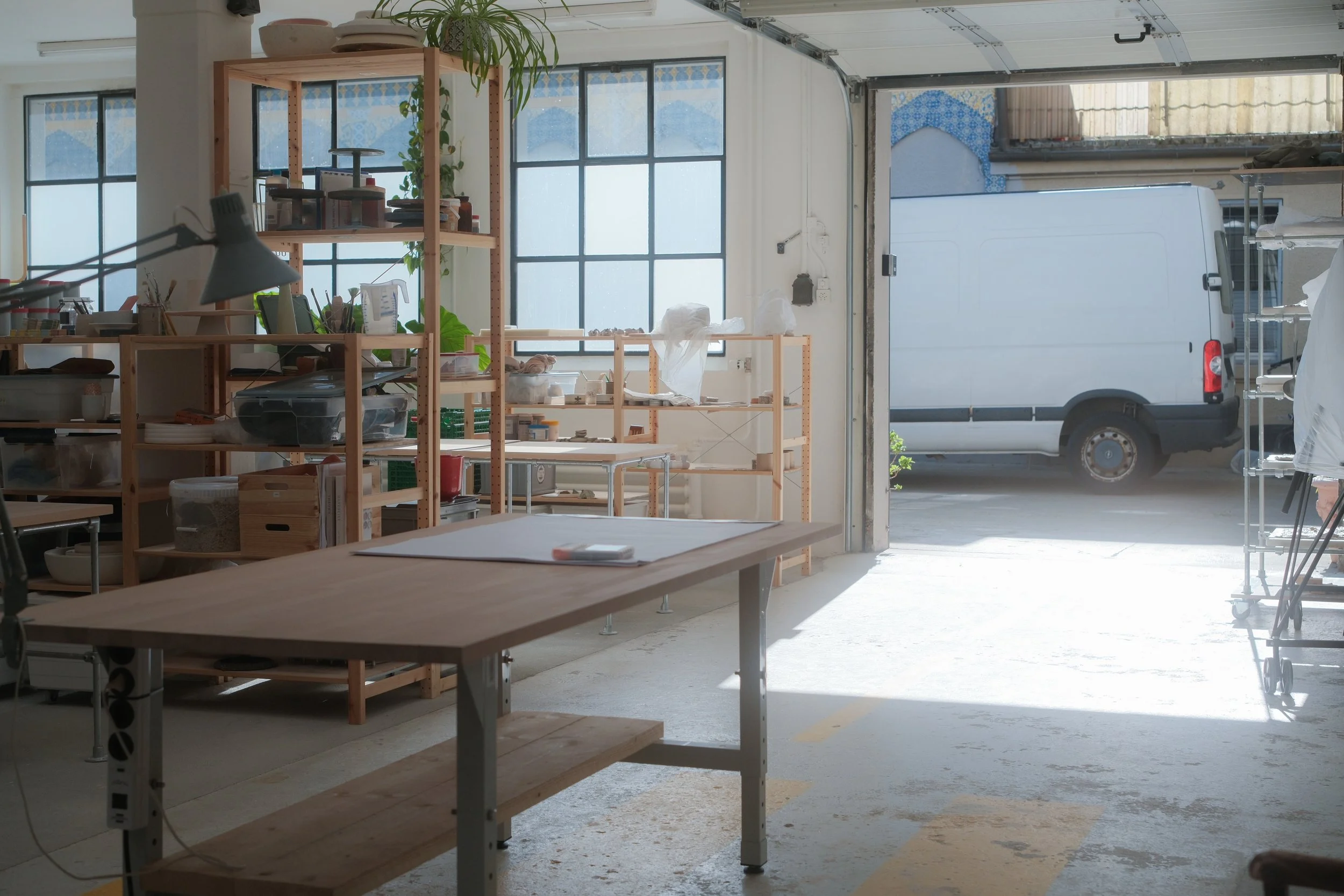 Ein Blick in eine Werkstatt oder einen Lagerraum mit großen Fenstern, Holzregalen, Arbeitstischen, Werkzeugen und einem weißen Lieferwagen vor der Tür.