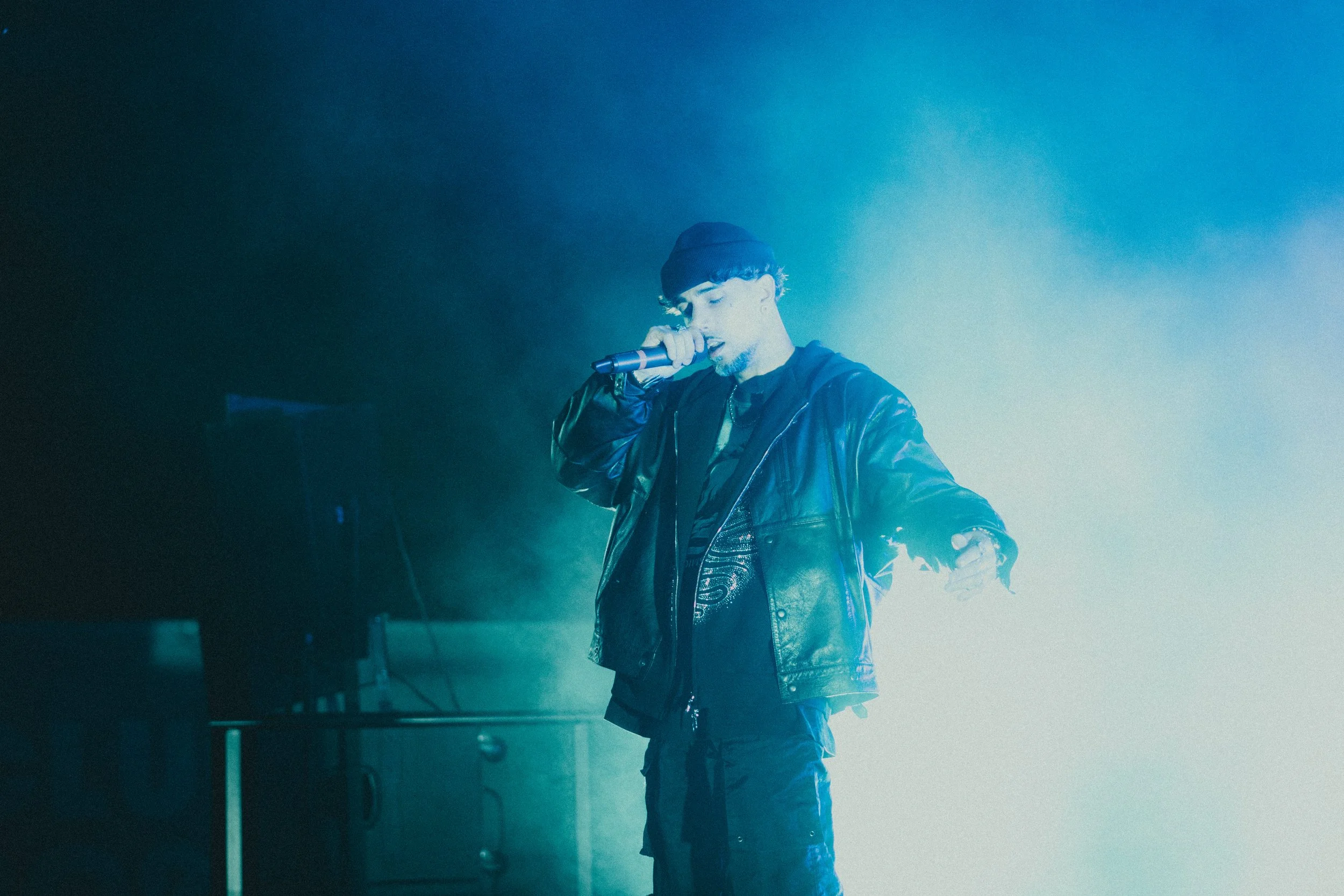 Un artiste chante en tenant un microphone, portant un blouson en cuir et un bonnet, dans une ambiance sombre éclairée de lumière bleue.