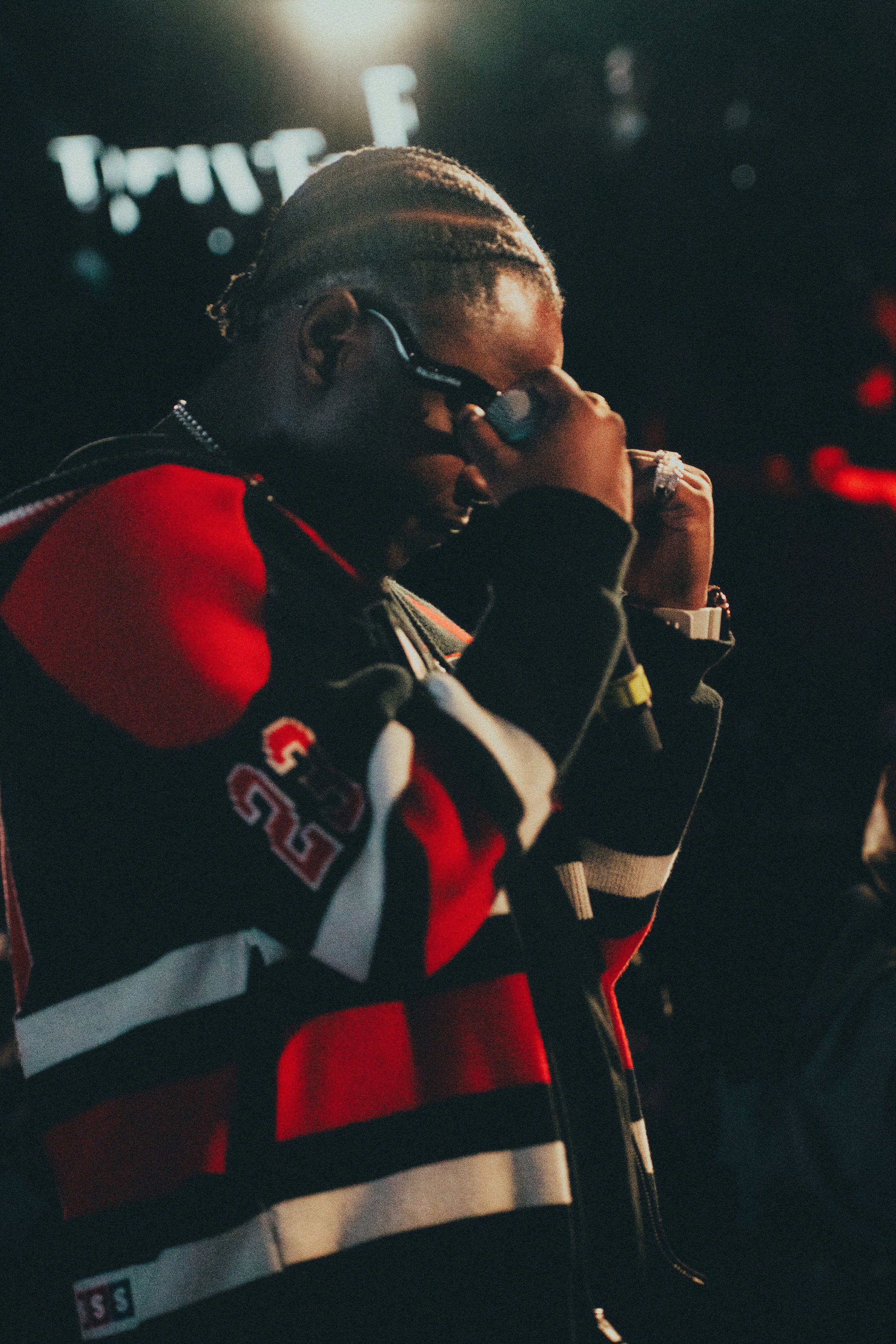 Un homme avec des cheveux tressés, portant des lunettes de soleil, un sweat à manches longues avec des couleurs noir, rouge, blanc et des numéros, semble en pleine réflexion ou prière dans un environnement sombre avec des lumières rougeâtres.