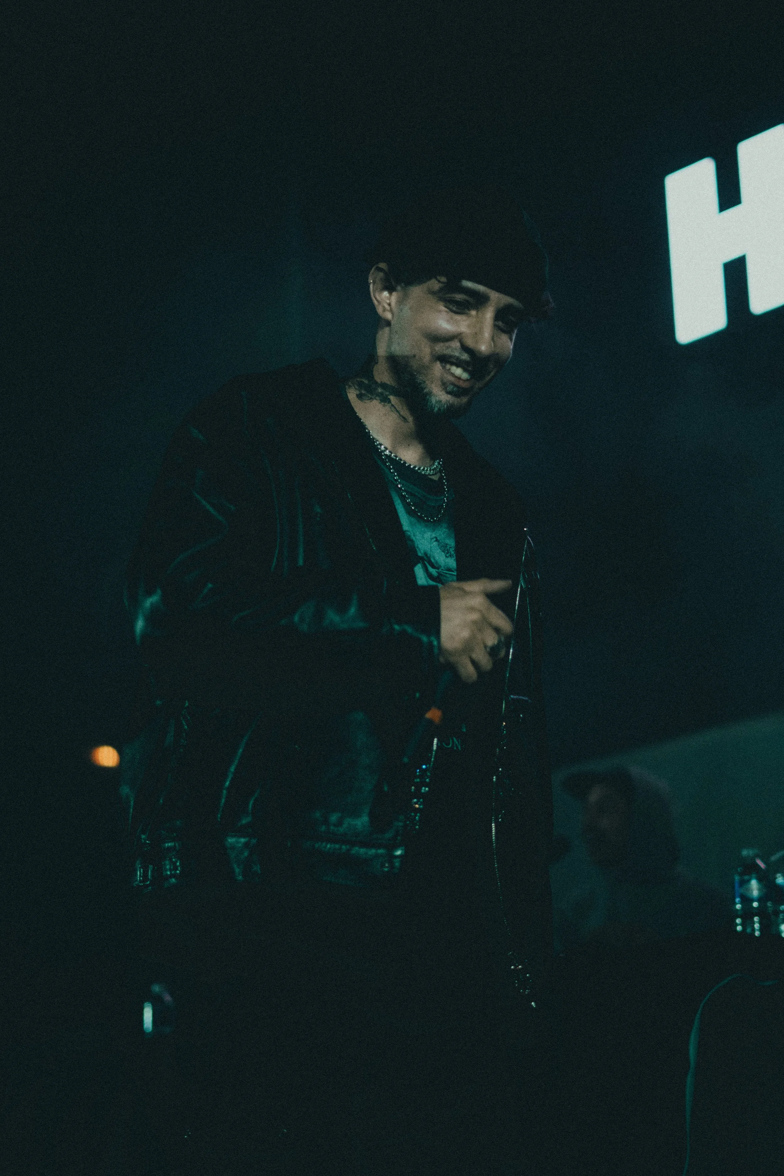 Un homme souriant tenant un microphone dans une ambiance sombre, éclairé par une lumière bleue, portant des chaînes et une veste noire, lors d'une performance ou d'un événement.