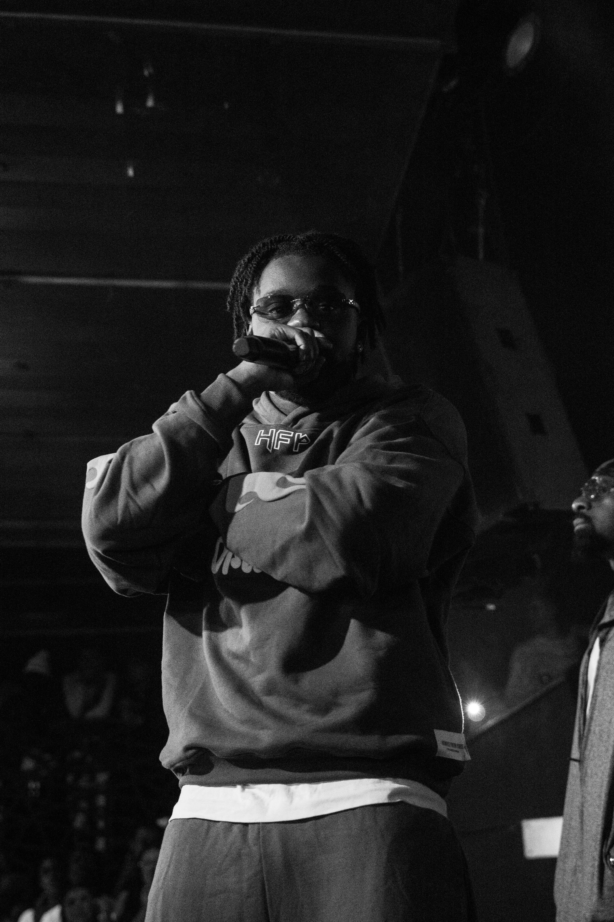Un jeune homme tenant un microphone, portant des lunettes, dans une scène en noir et blanc, probablement lors d'une performance ou d'un événement en intérieur.