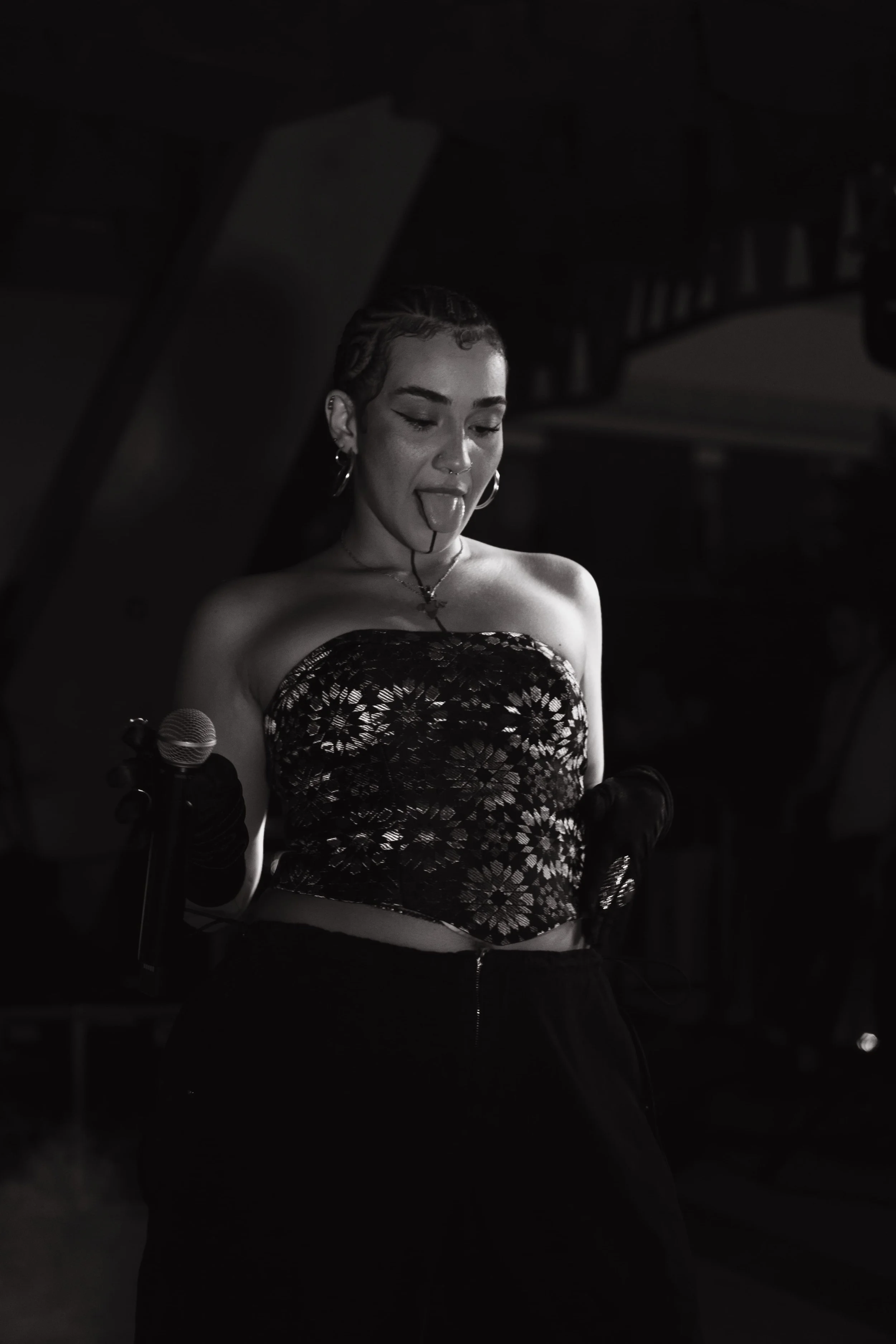 Une femme jeune avec des cheveux courts bouclés, portant un haut en dentelle, tenant un microphone, en train de faire une grimace en tirant la langue, dans un environnement sombre