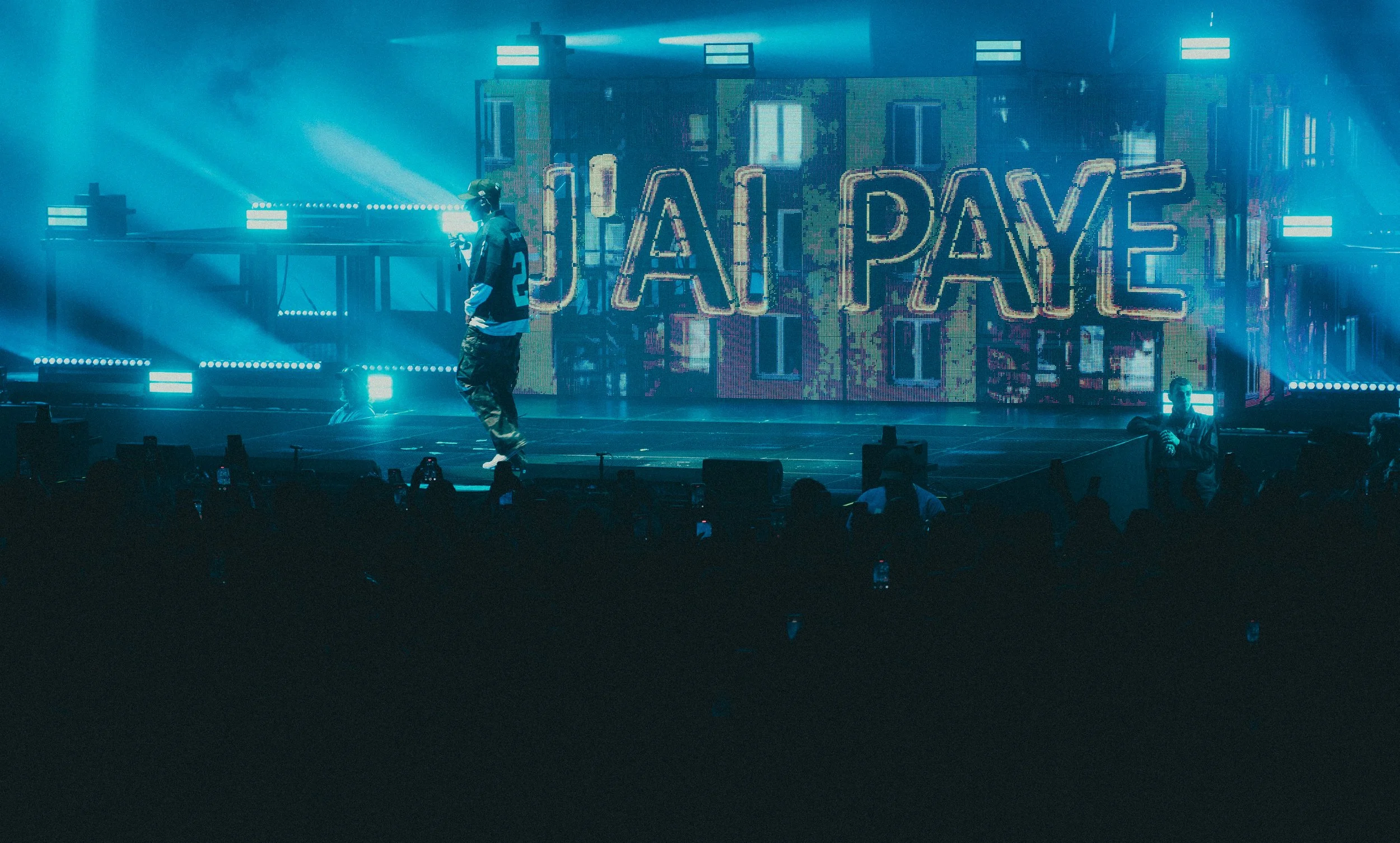 Un performer sur scène avec un écran géant en arrière-plan affichant le mot "UHAI PAYE" en lettres lumineuses, entouré de lumières bleues et de fumée, avec un public regardant.