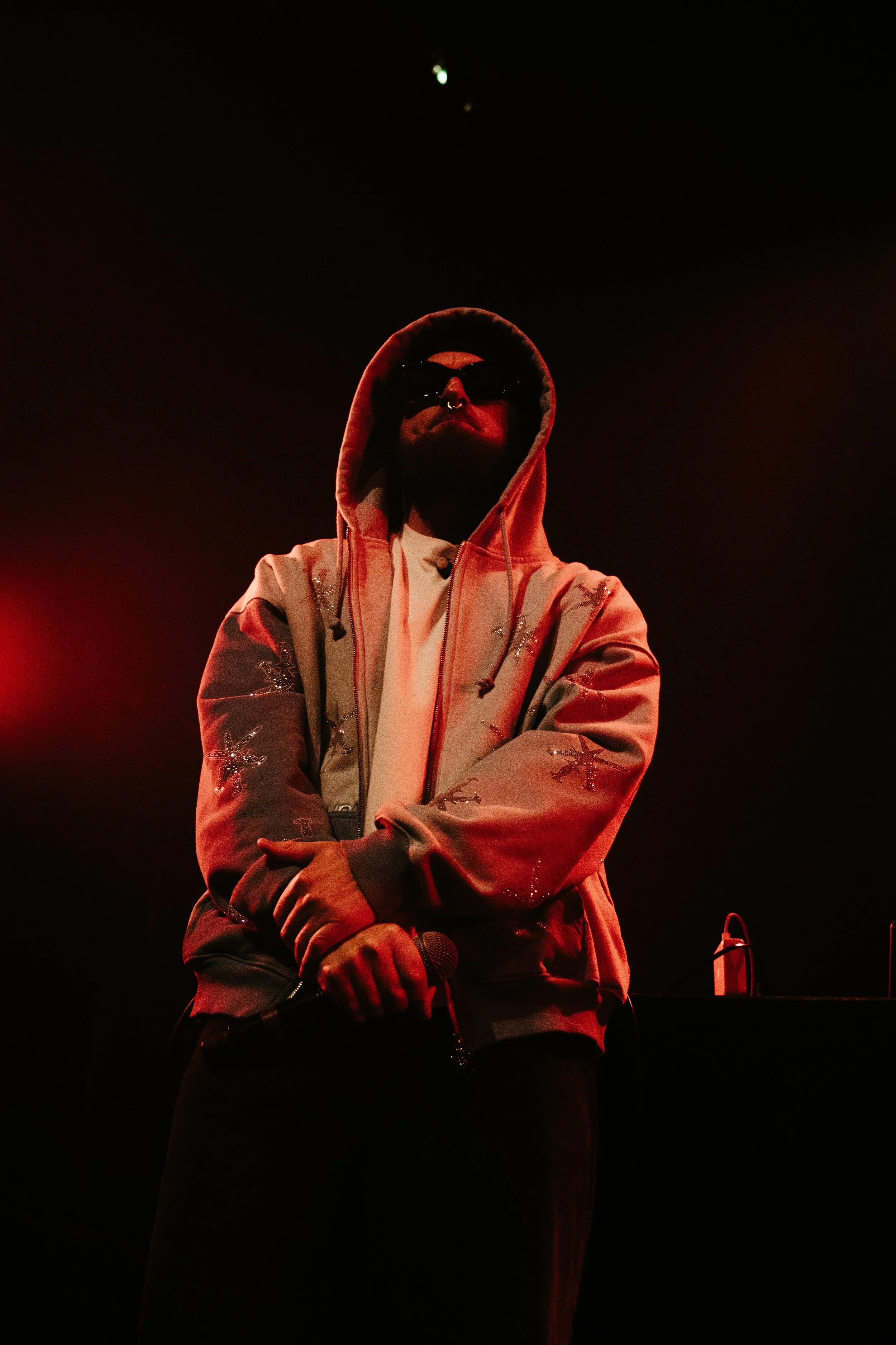Un homme portant un sweat à capuche avec des motifs brillants, des lunettes de soleil noires, tenant un microphone dans une main, sous un éclairage rouge en scène.