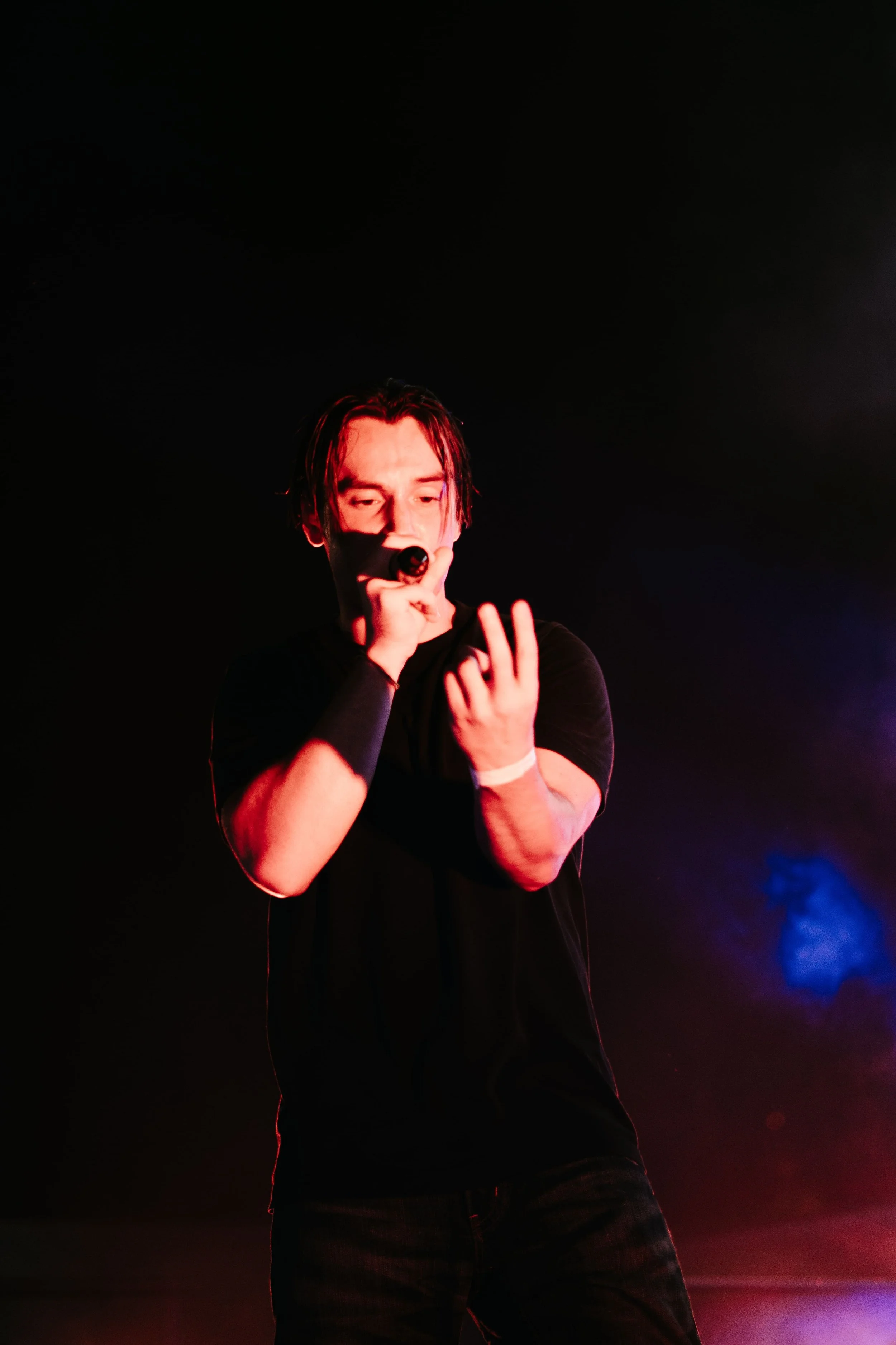 Un jeune homme avec les cheveux longs tient un microphone lors d'une performance sur scène, avec un fond sombre et des lumières colorées