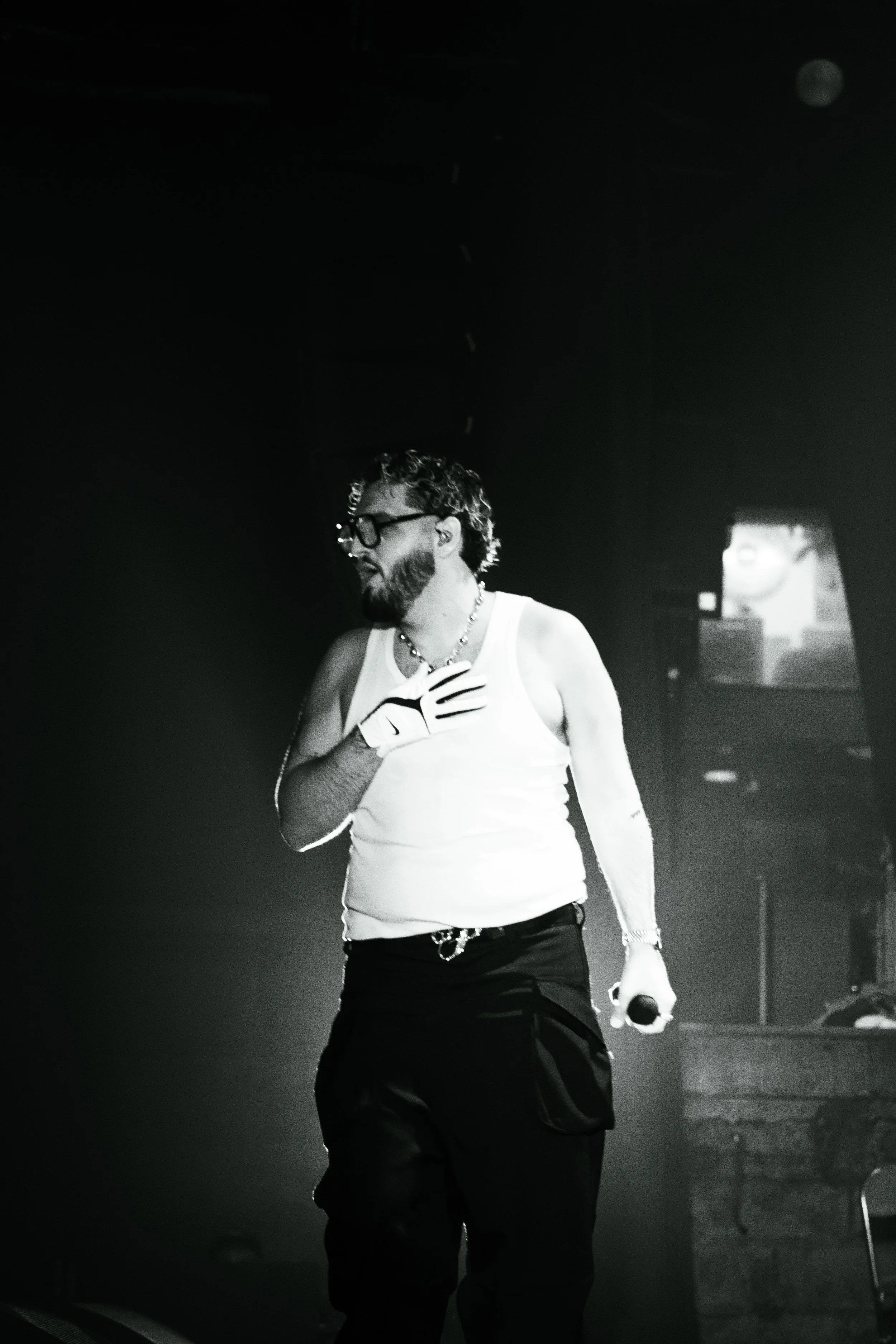 Un homme porte un t-shirt blanc, des lunettes et une chaîne, tient un microphone dans la main, face à une scène sombre.