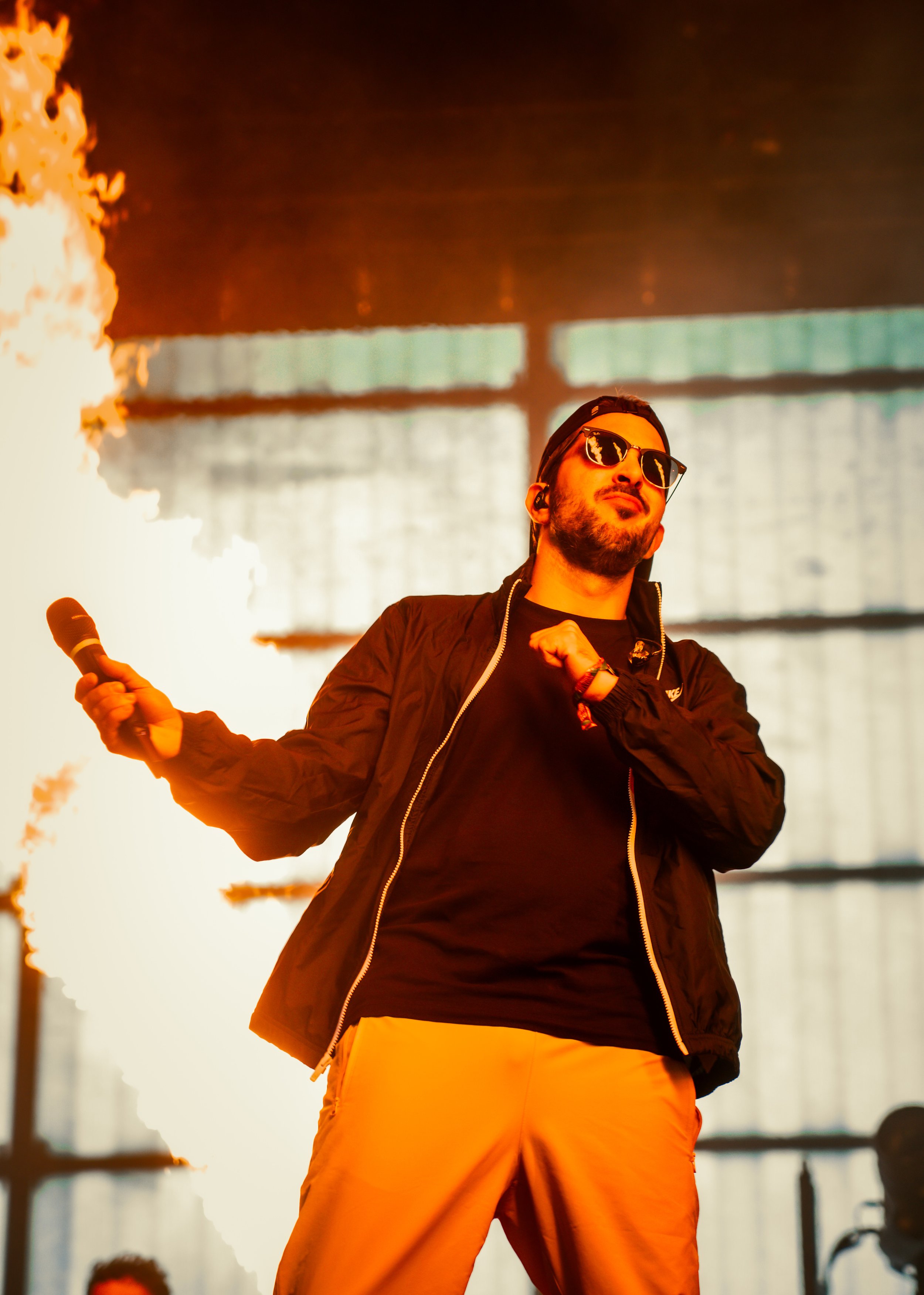 Un homme noir portant des lunettes de soleil, une veste noire, un t-shirt noir, et un pantalon jaune, tenant un microphone, sur scène avec un feu d'artifice en arrière-plan.