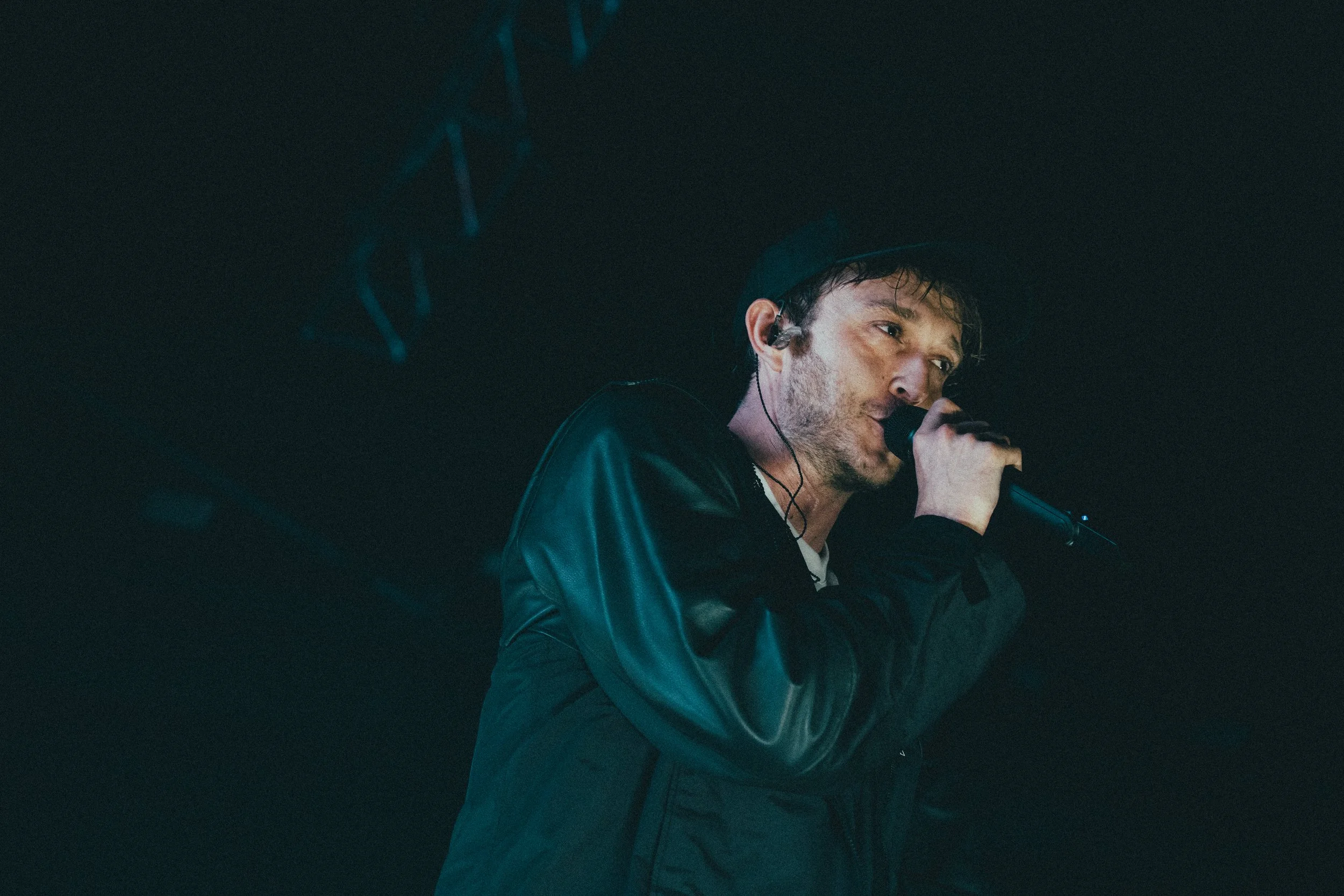 Un homme chante dans un micro lors d'une performance, portant une veste en cuir noire et un chapeau noir, avec un éclairage sombre et bleu.