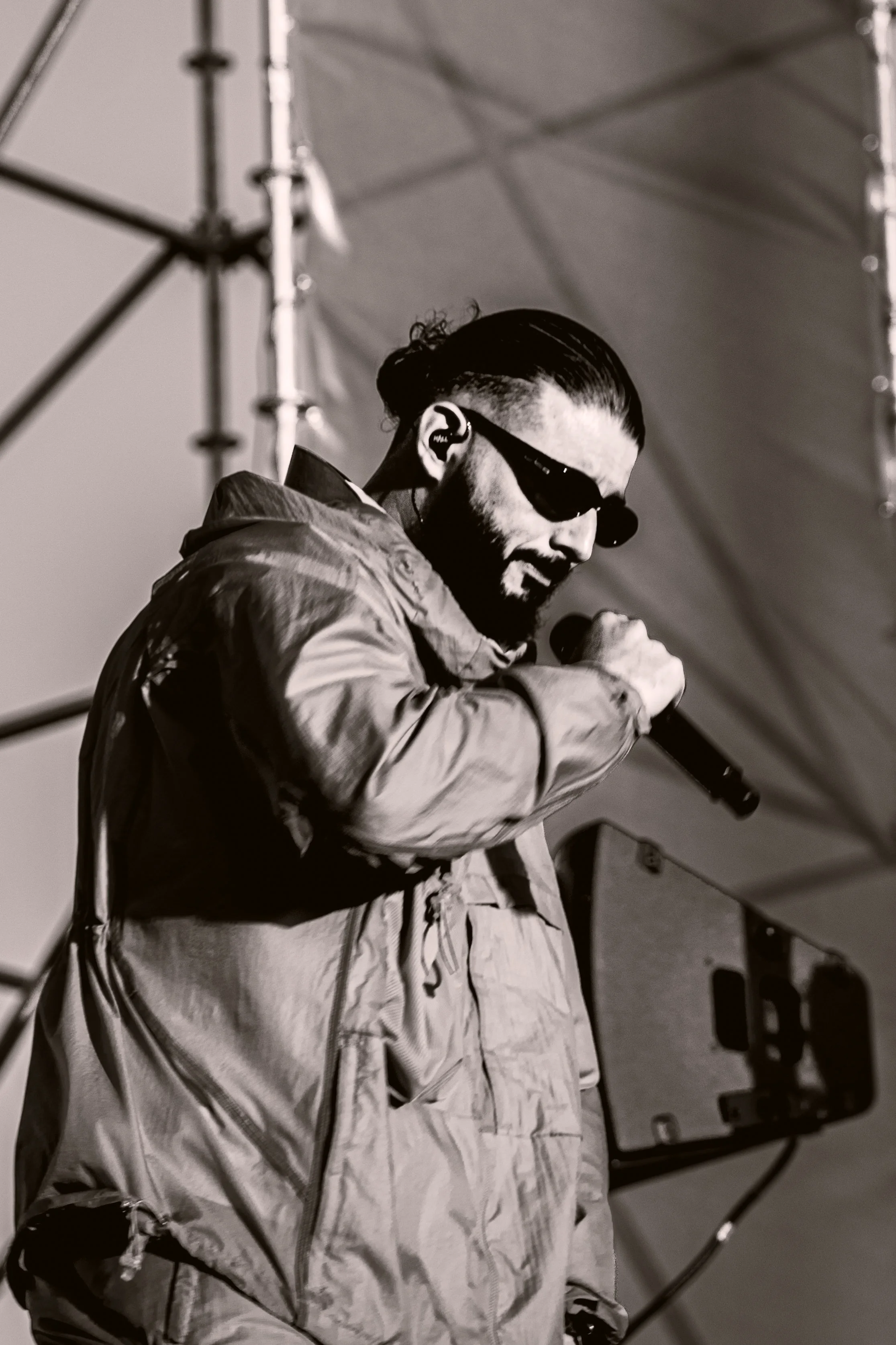 Un homme portant des lunettes de soleil noires, un casque d'écoute, une barbe, une moustache et une veste ample, tenant un microphone dans une main, semble être en train de chanter ou de parler sur scène