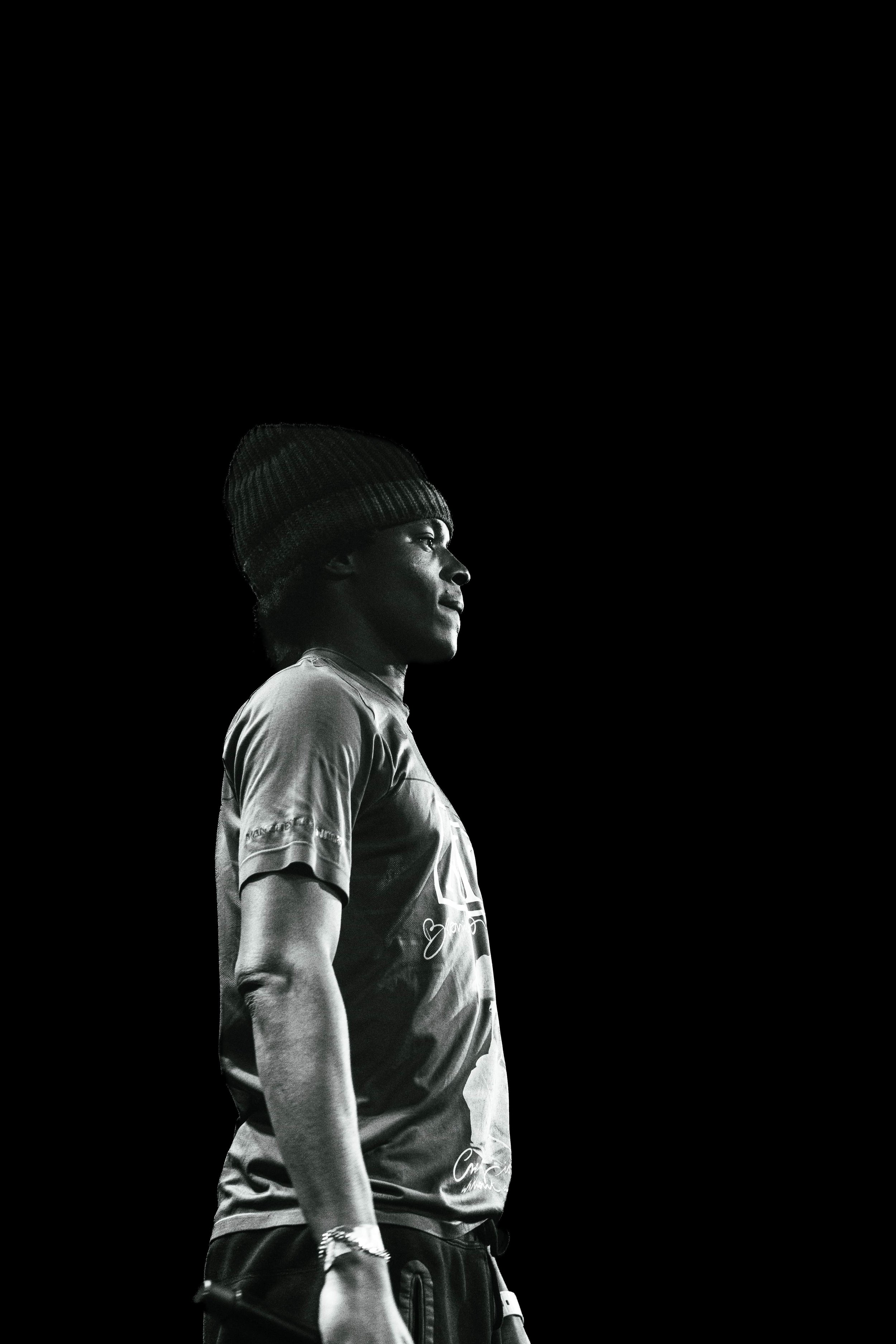 Portrait en noir et blanc d'un homme portant une casquette en laine, vêtu d'un t-shirt, regardant vers la droite, avec un fond noir.