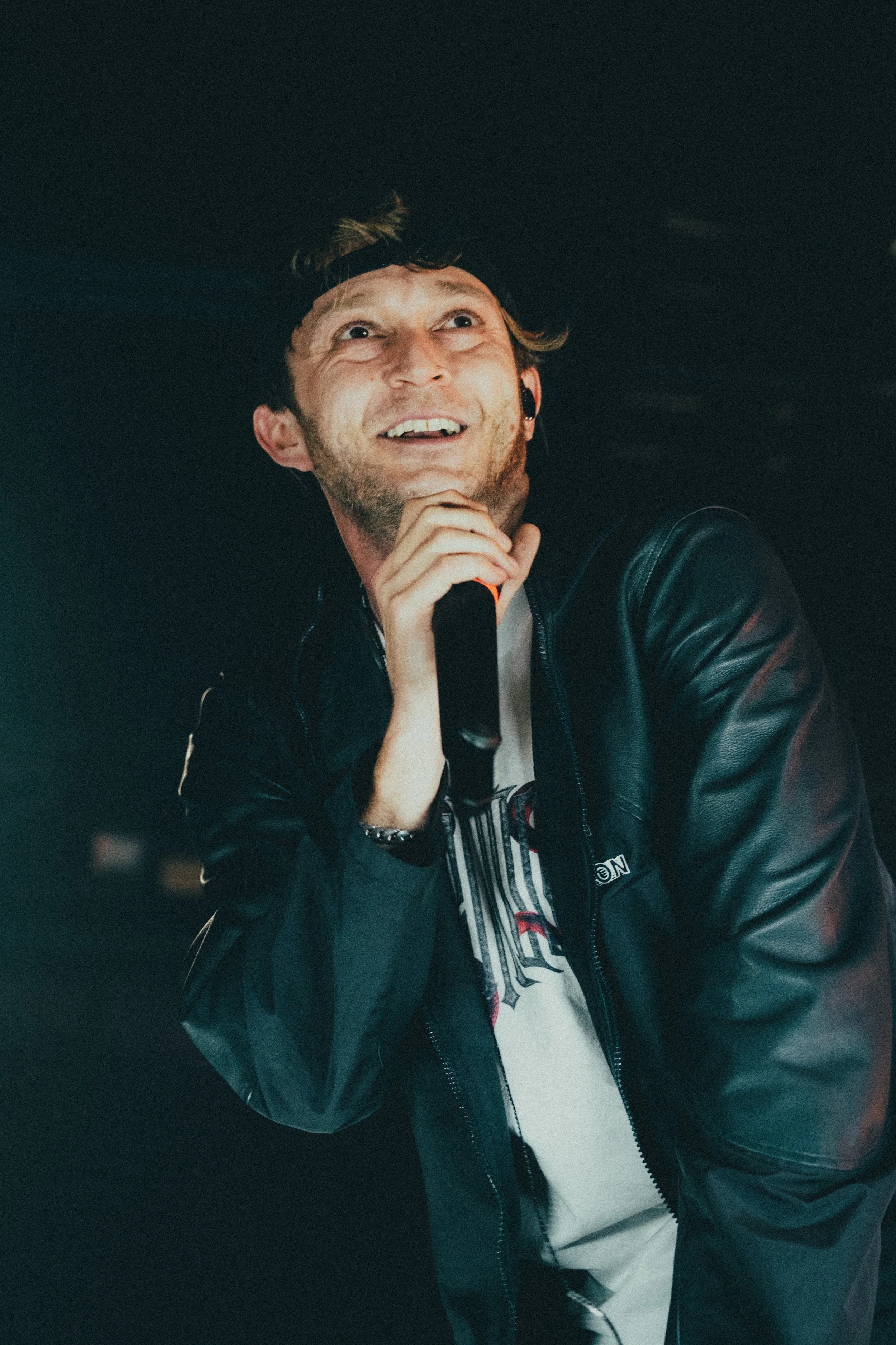Un homme qui chante ou parle dans un microphone, portant une veste en cuir noir et un T-shirt blanc, souriant dans une ambiance sombre.