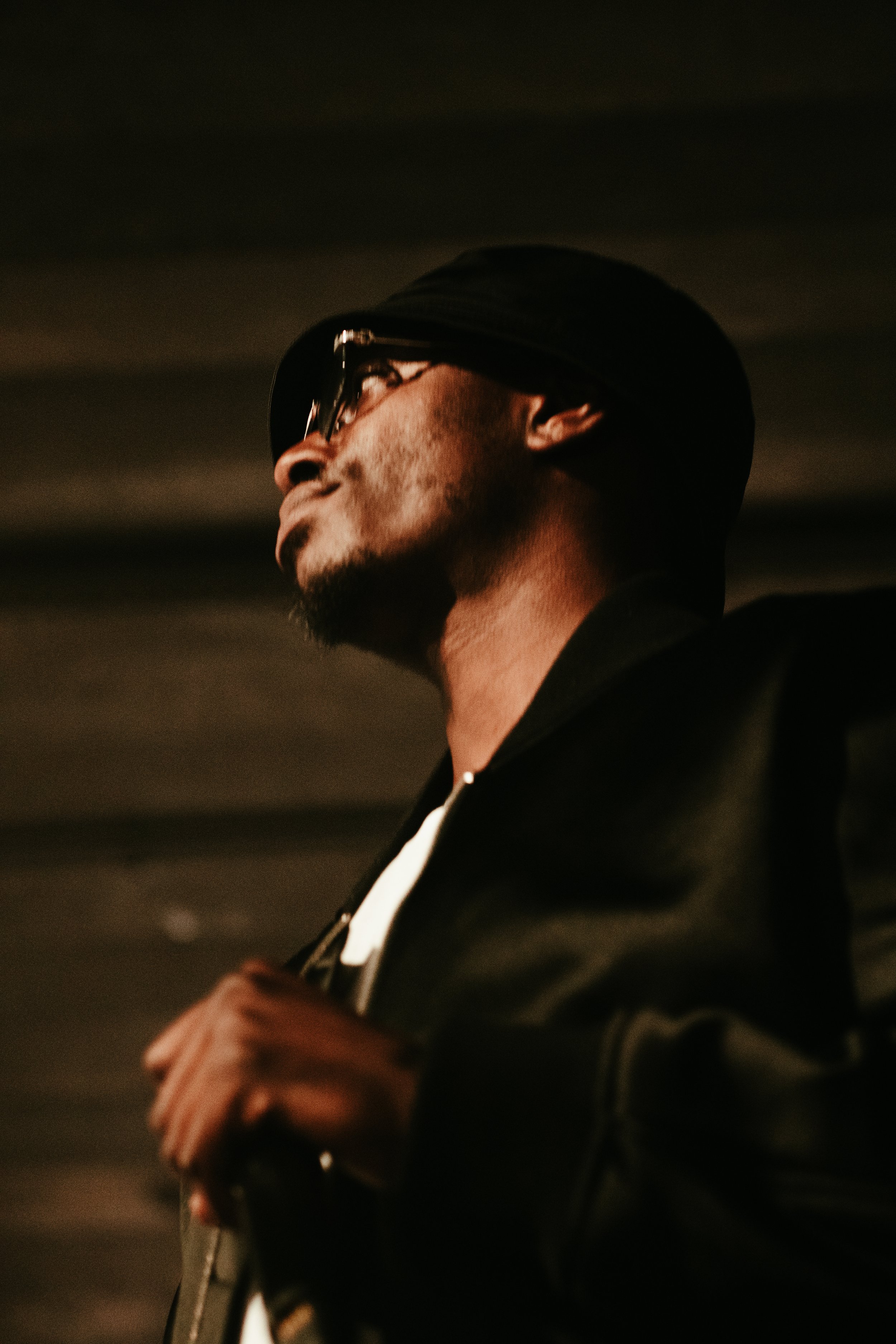 Homme portant une casquette noire, lunettes sombre, en tenu noir, vue de profil dans un environnement sombre.