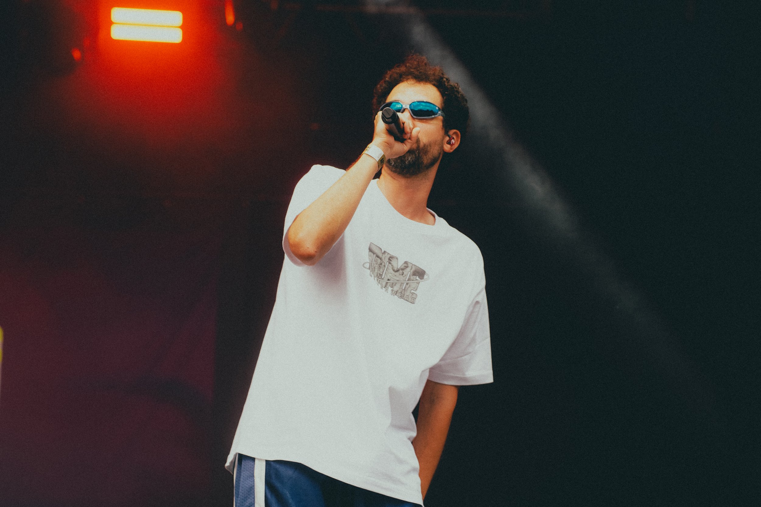 Un homme portant des lunettes de soleil andiennes, un t-shirt blanc avec logo, tient un microphone en scène avec éclairage rouge et noir en arrière-plan.