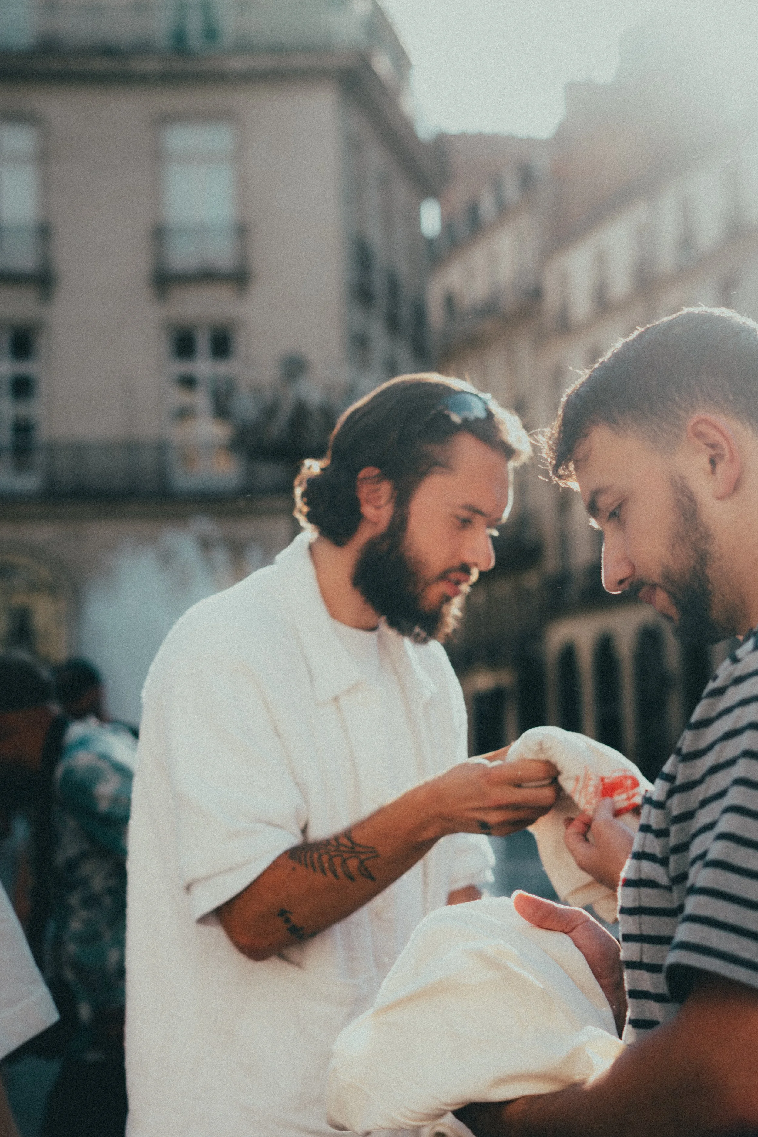 Deux hommes discutent dans une rue ensoleillée, l'un portant une chemise blanche et l'autre une chemise rayée.