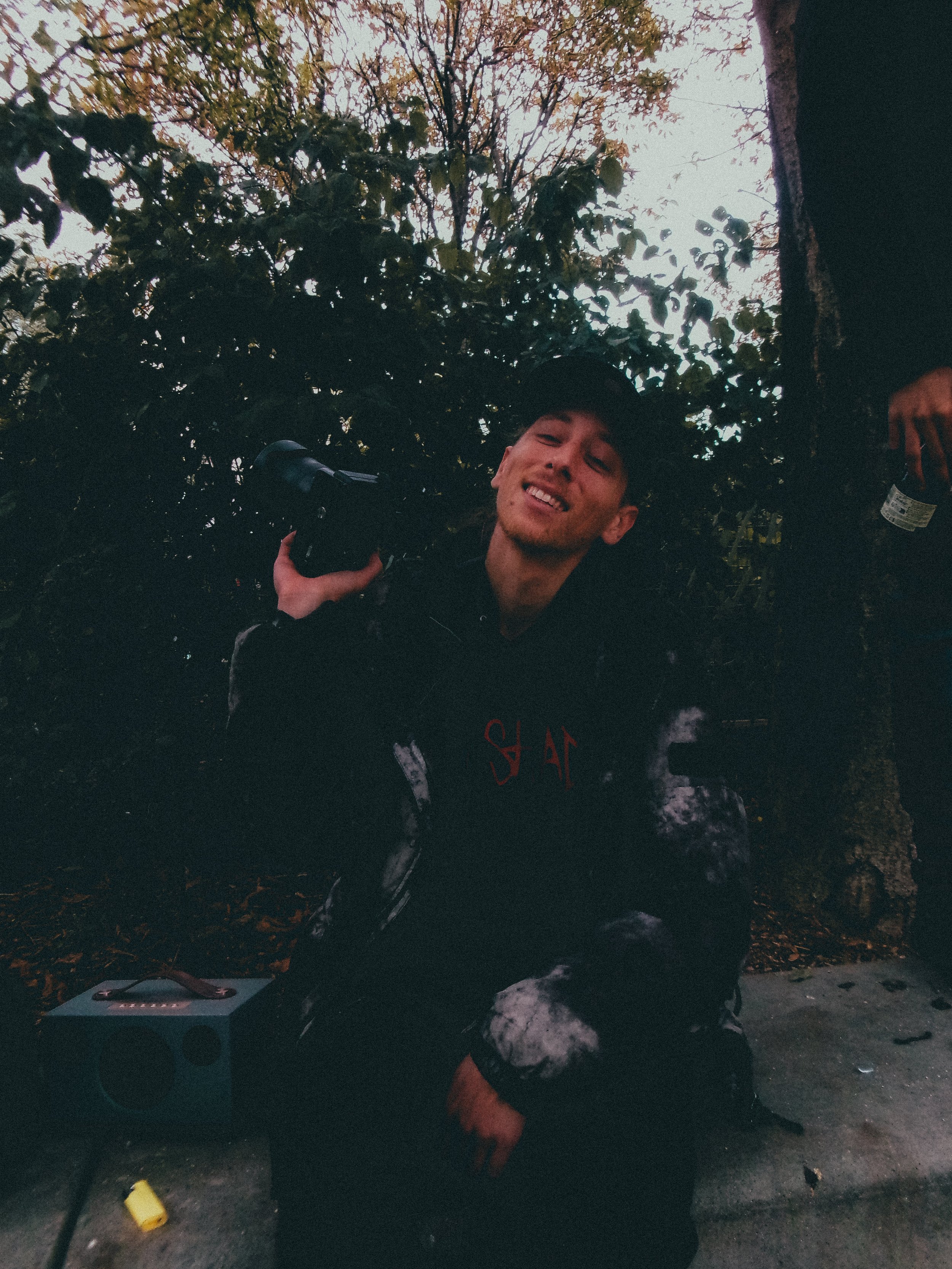 Jeune homme souriant, tenant un appareil photo, assis devant un arbre, dans un environnement extérieur forestier.
