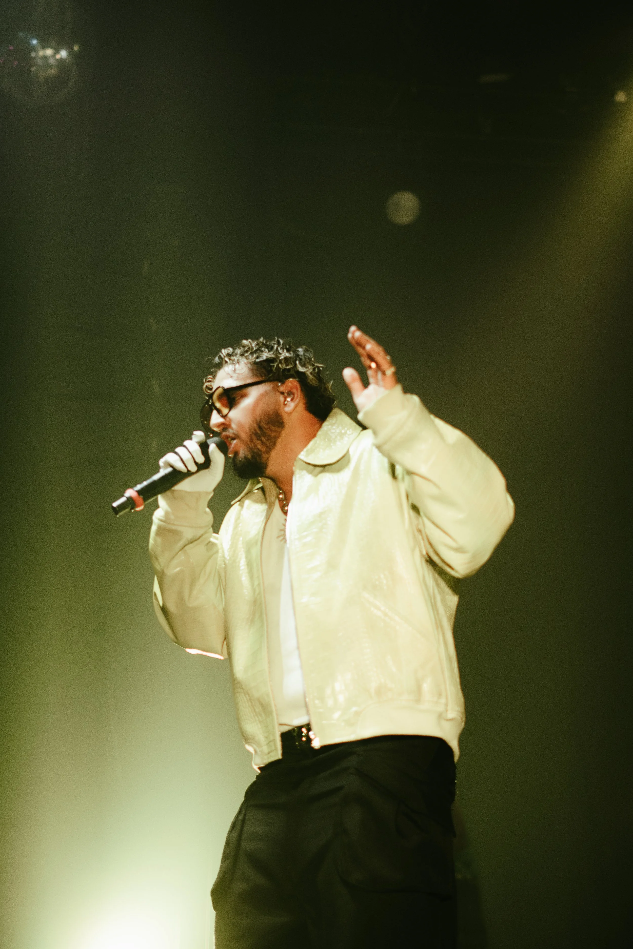 Un homme avec des lunettes noires chante en tenant un microphone lors d'une performance sur scène, vêtu d'une veste beige lumineux et de pantalons sombres, sous un éclairage de scène vert et jaune.