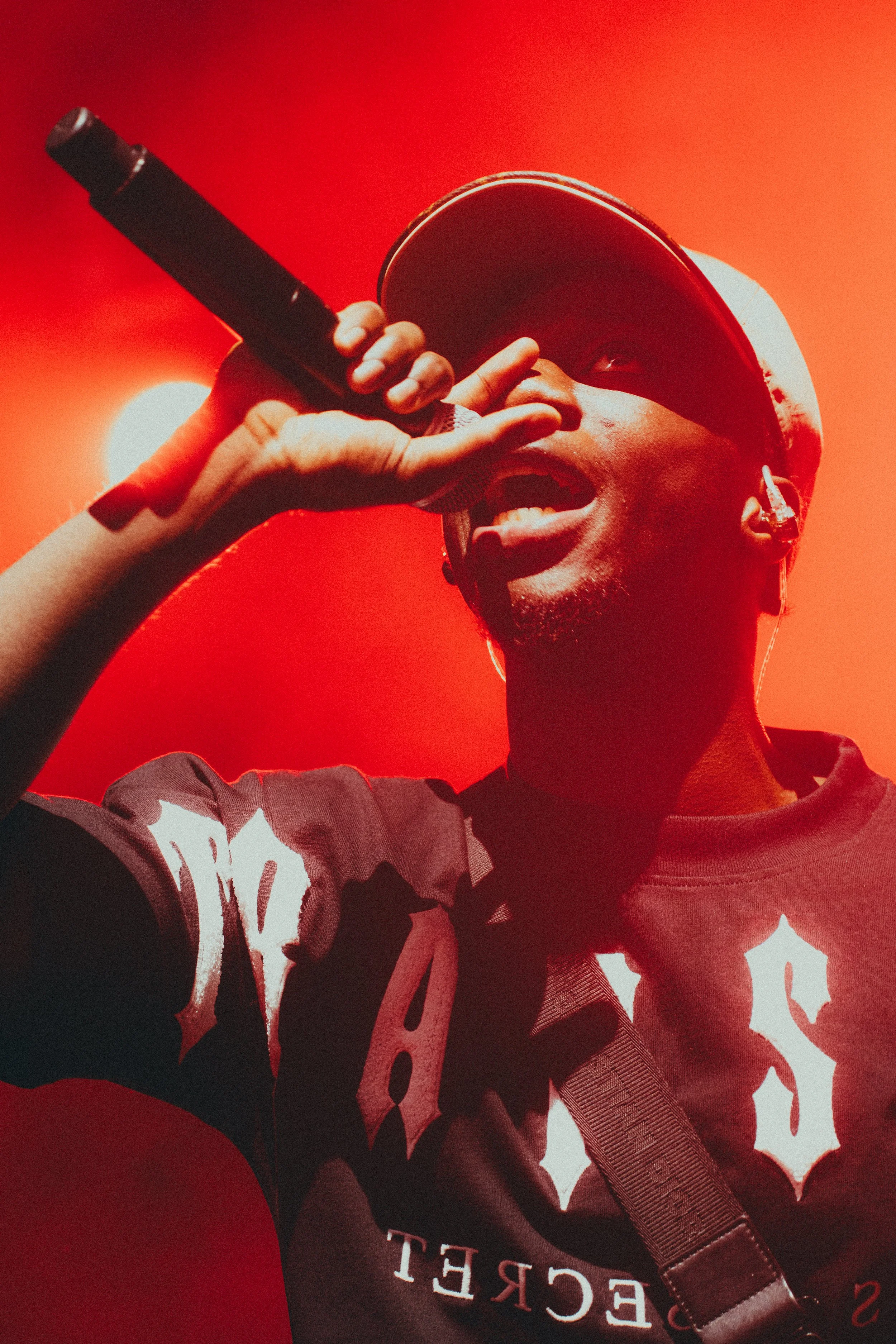Un jeune homme portant une casquette et un bonnet, tenant un micro, semblant en train de rapper ou de chanter, avec un fond rouge vif.