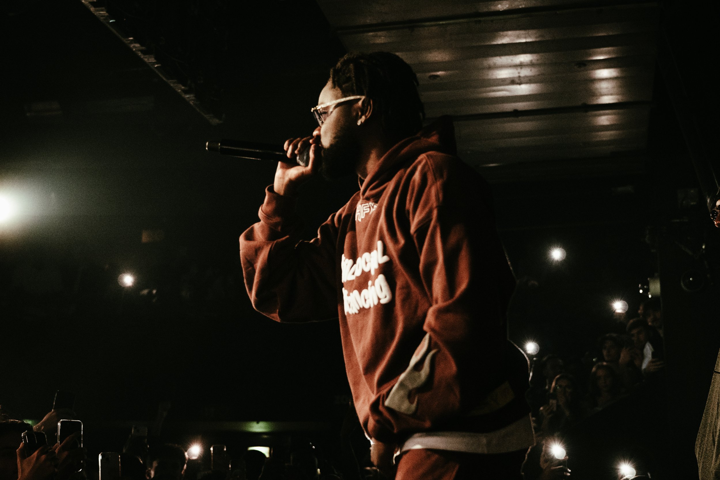 Un artiste ou rappeur portant une veste marron et des lunettes, tenant un micro, en train de performer sur scène pendant un concert.