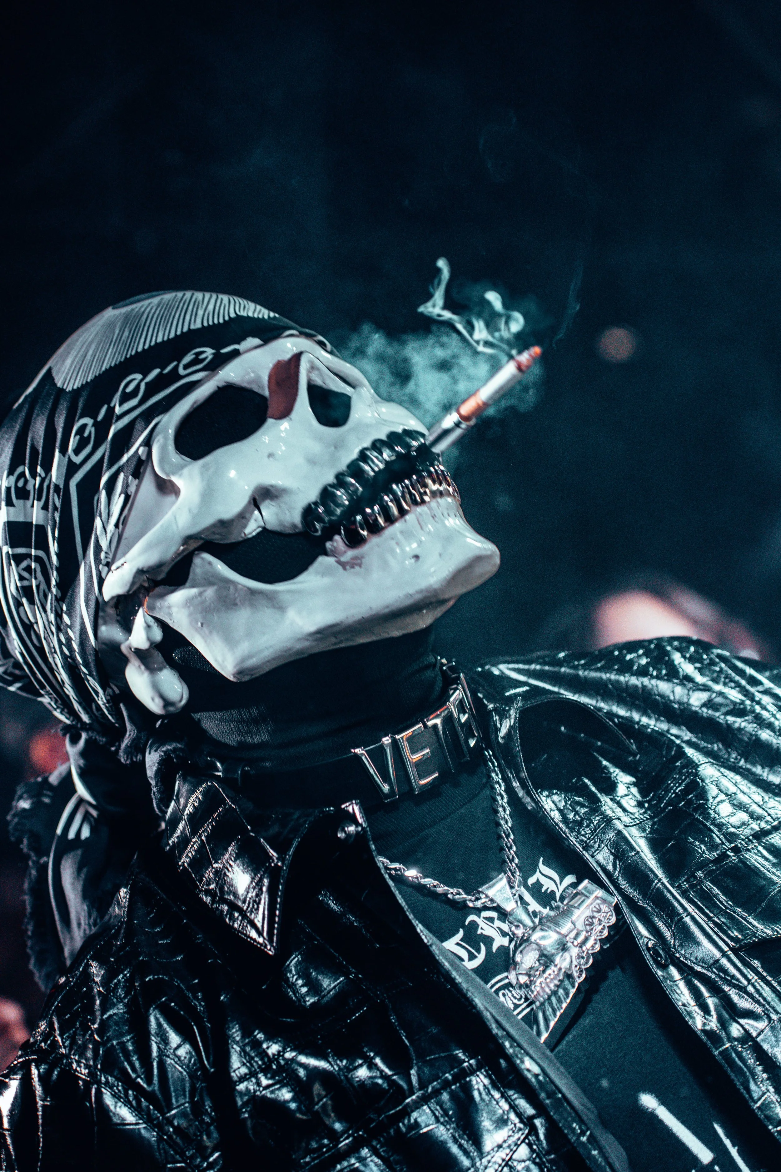 Une personne porte un masque de squelette blanc, un bandana noir et blanc, une veste en cuir noir, un collier avec un pendentif tête de mort, et un collier avec le mot 'VET'. Elle fume une cigarette, avec de la fumée qui s'élève dans l'air.