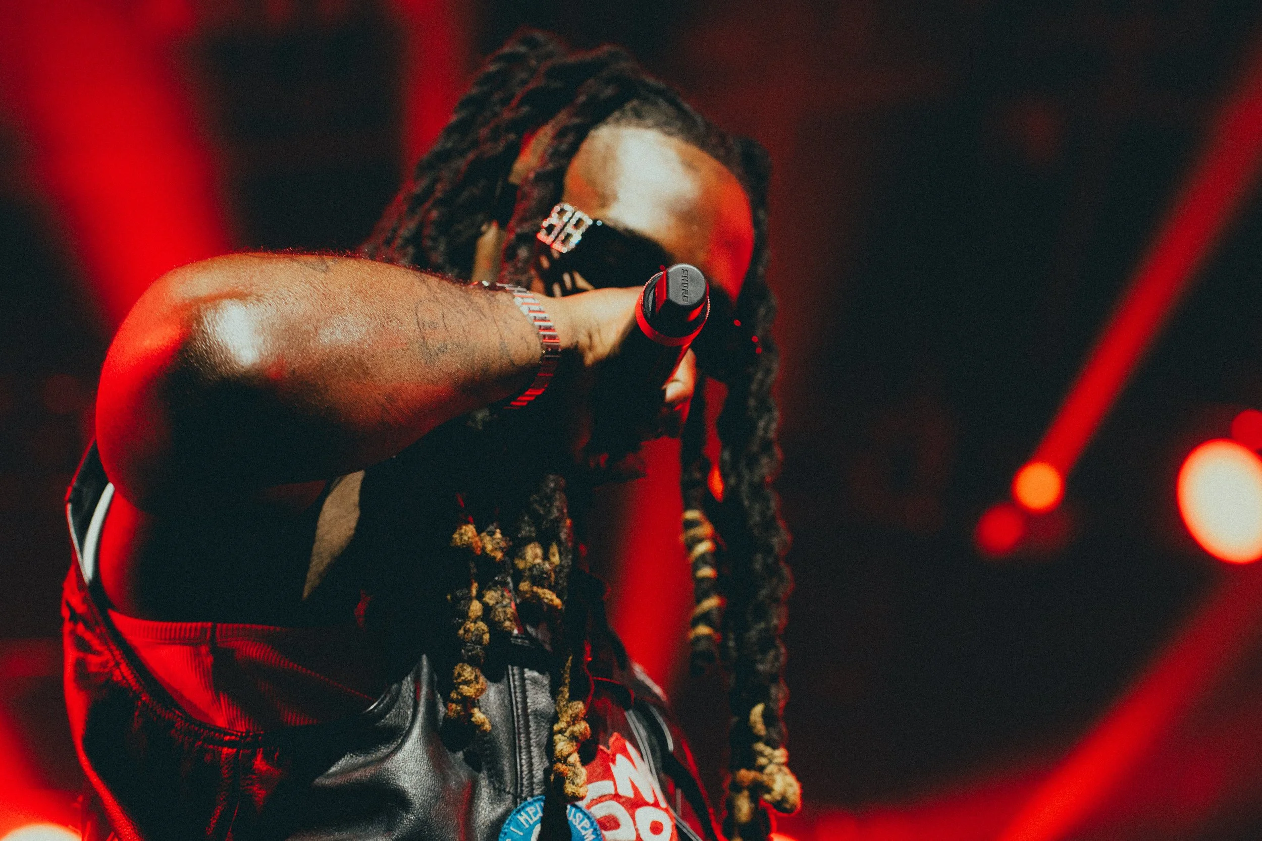Rappeur avec dreadlocks portant des lunettes de soleil, un hoodie noir et un gilet en cuir, tenant un microphone et performant sur scène avec des lumières rouges en arrière-plan.