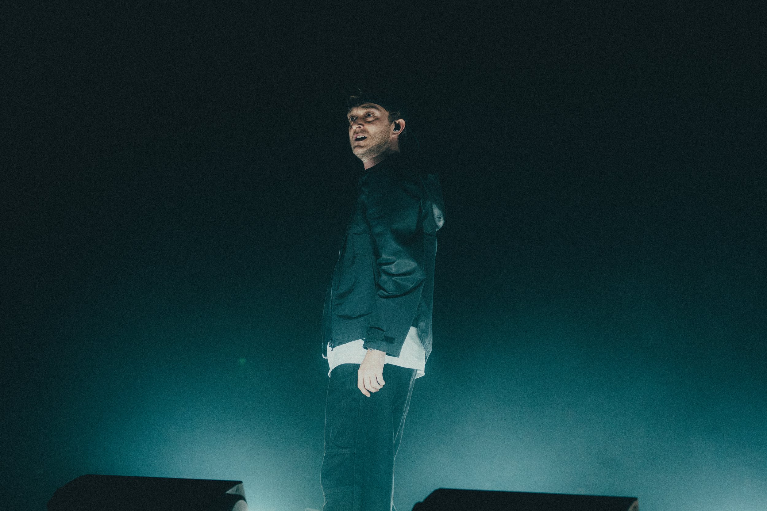 Un homme debout sur scène, portant une veste noire, avec un arrière-plan sombre et un éclairage tamisé