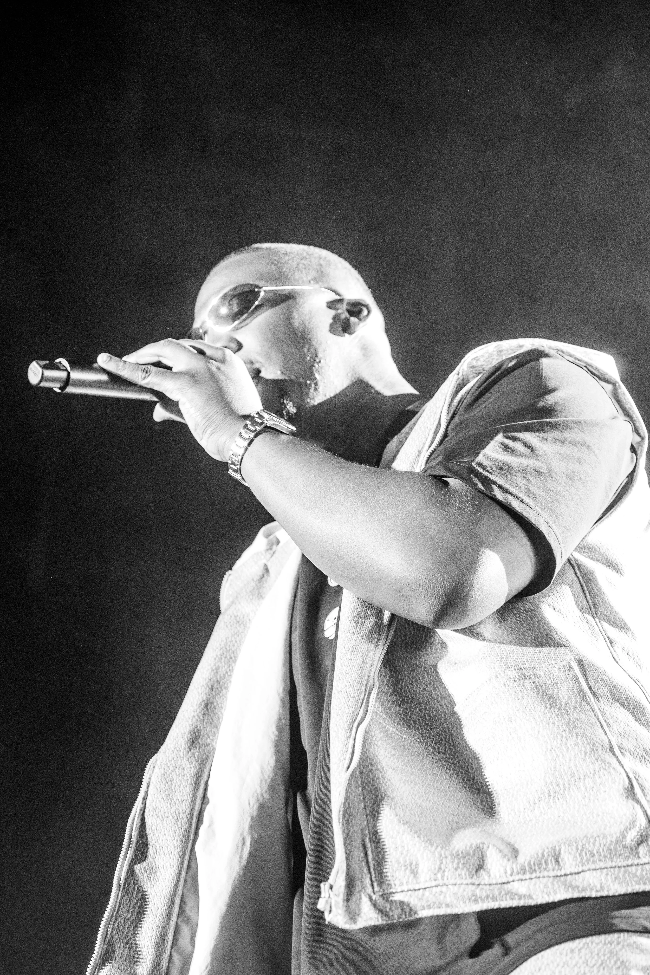Un homme portant des lunettes de soleil, une montre, et un vêtement sans manches, tenant un microphone, en train de performer sur scène en noir et blanc.