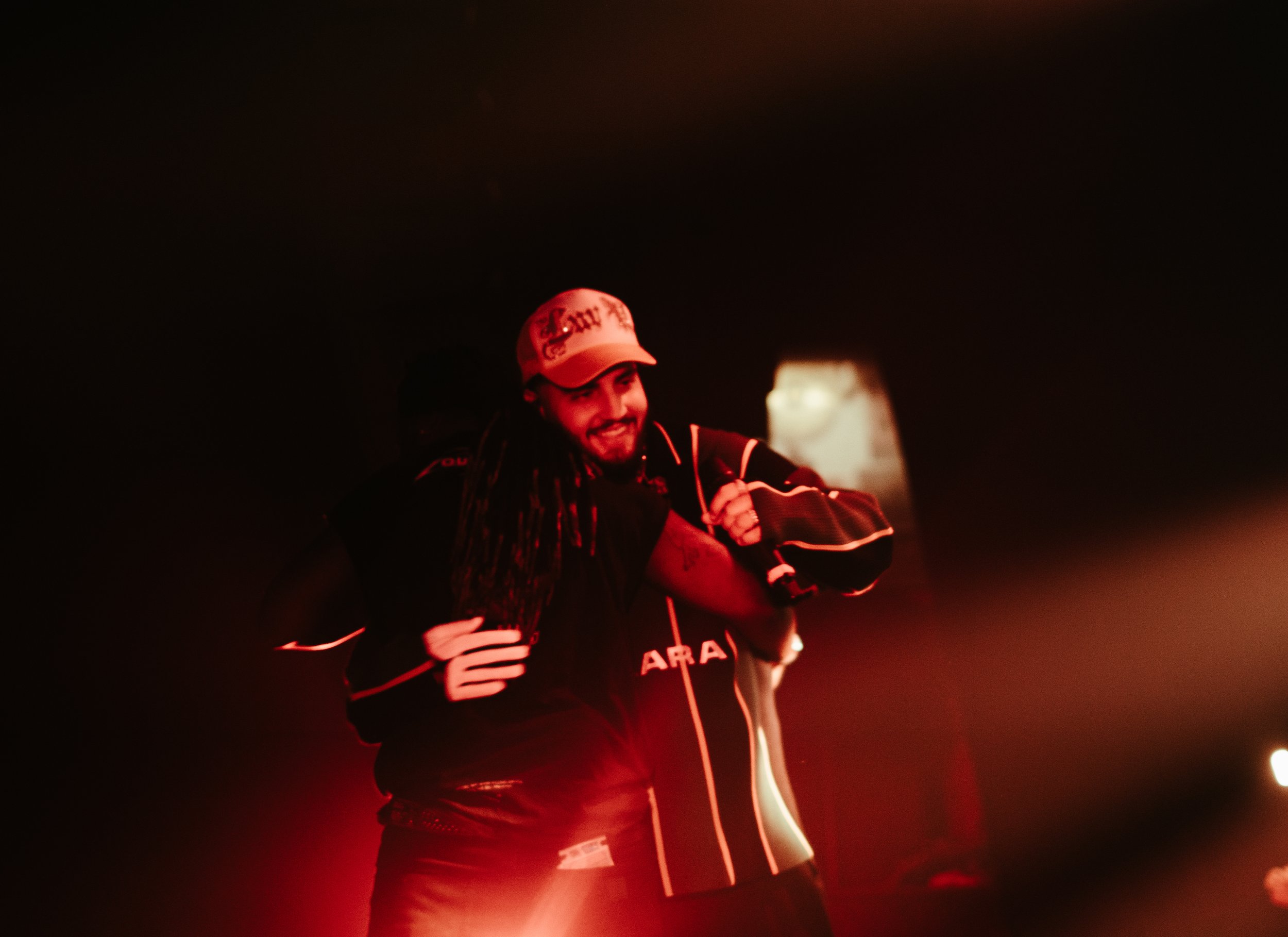 Une personne souriante, portant une casquette et une veste de sport, semble en train d'être félicitée ou de célébrer dans un environnement sombre, avec une lumière rougeâtre.
