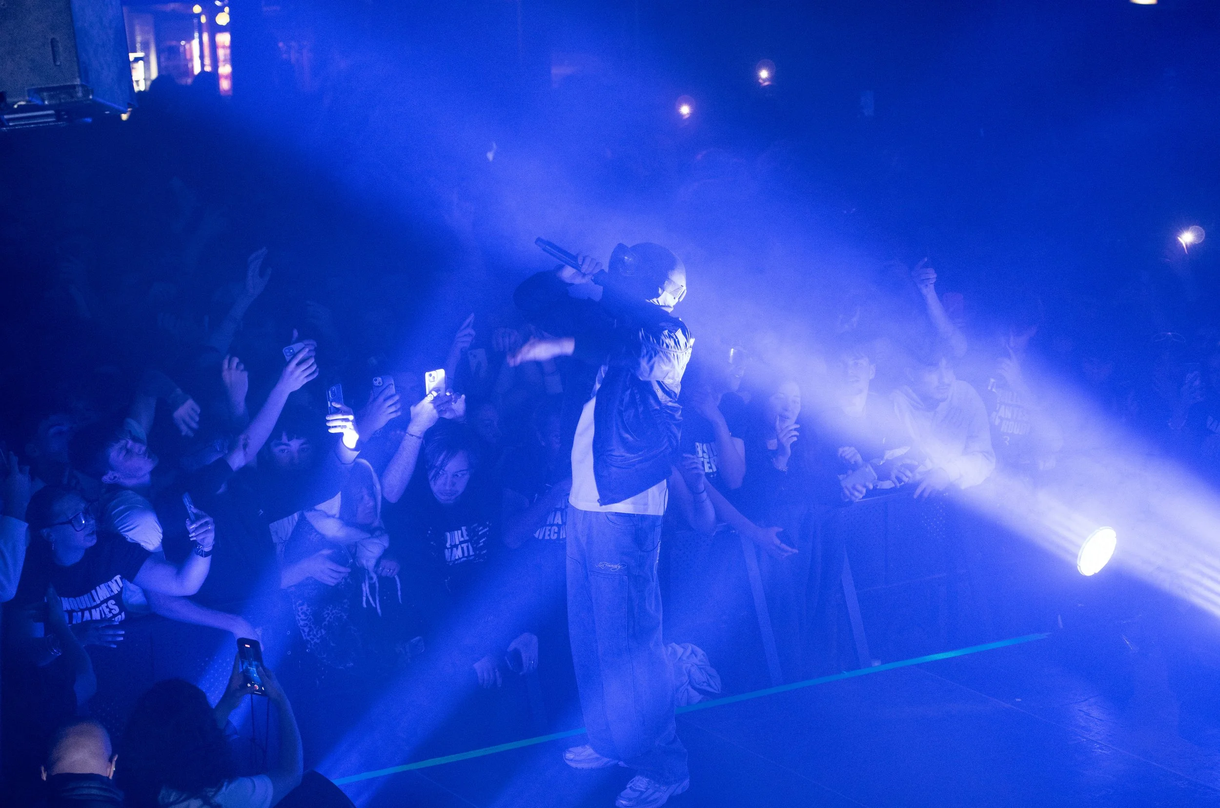 Un rappeur sur scène avec un micro, entouré d'une foule enthousiaste dans une lumière bleue.
