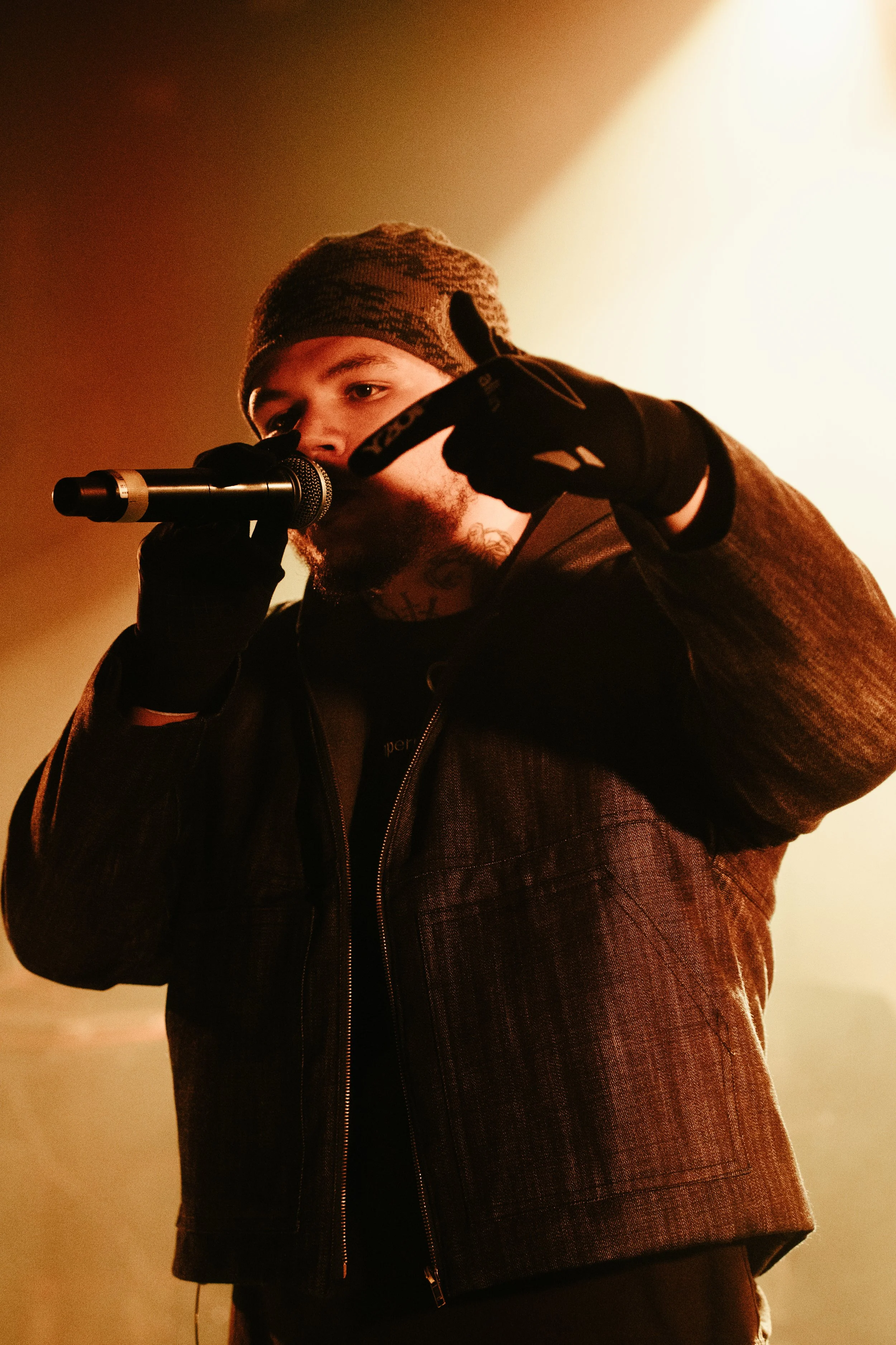 Un jeune homme portant un bonnet, une veste foncée, tenant un microphone, est sous un éclairage chaud.